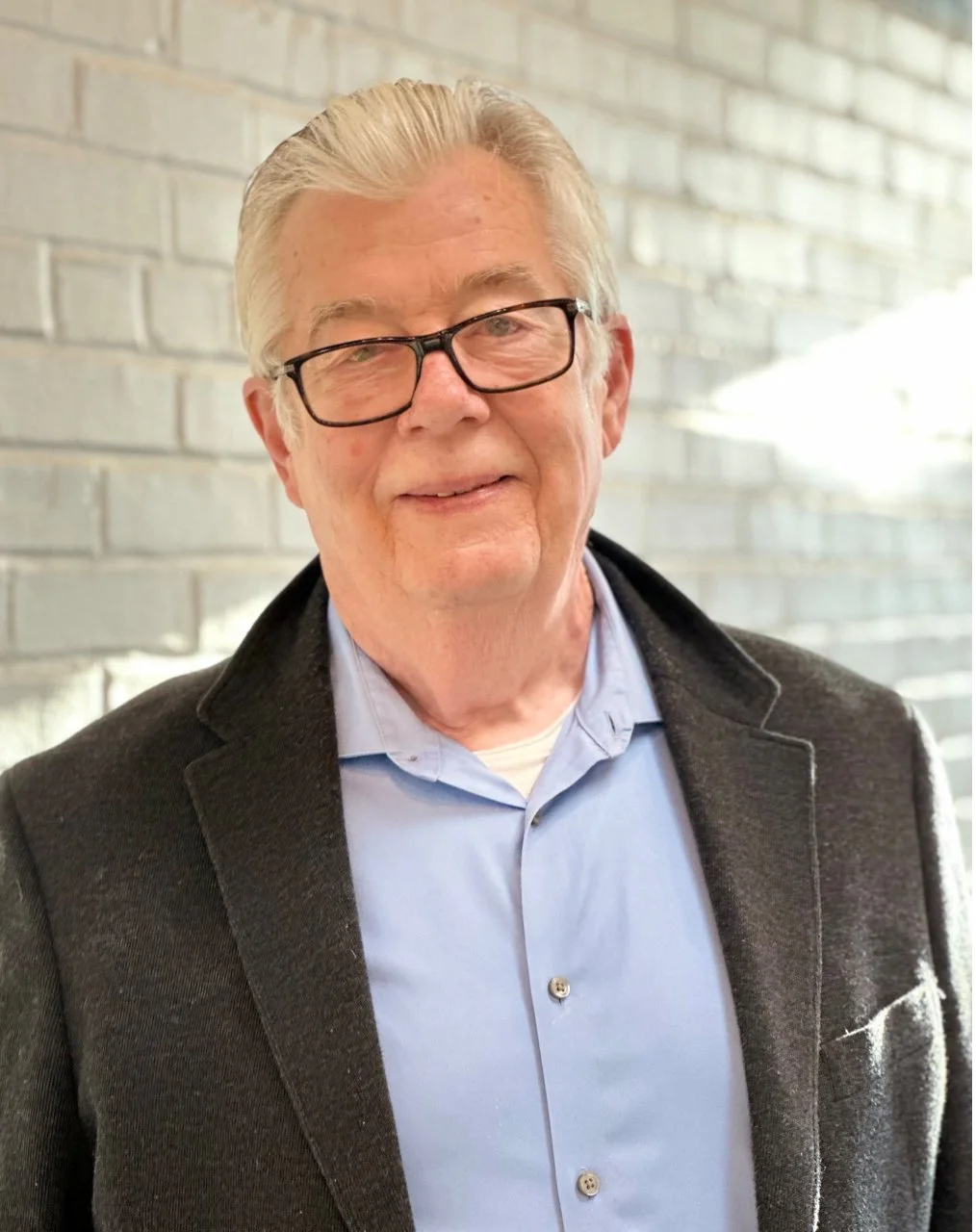 An elderly man with gray hair and glasses, smiling, wearing a light blue collared shirt and a dark blazer, standing in front of a brick wall.