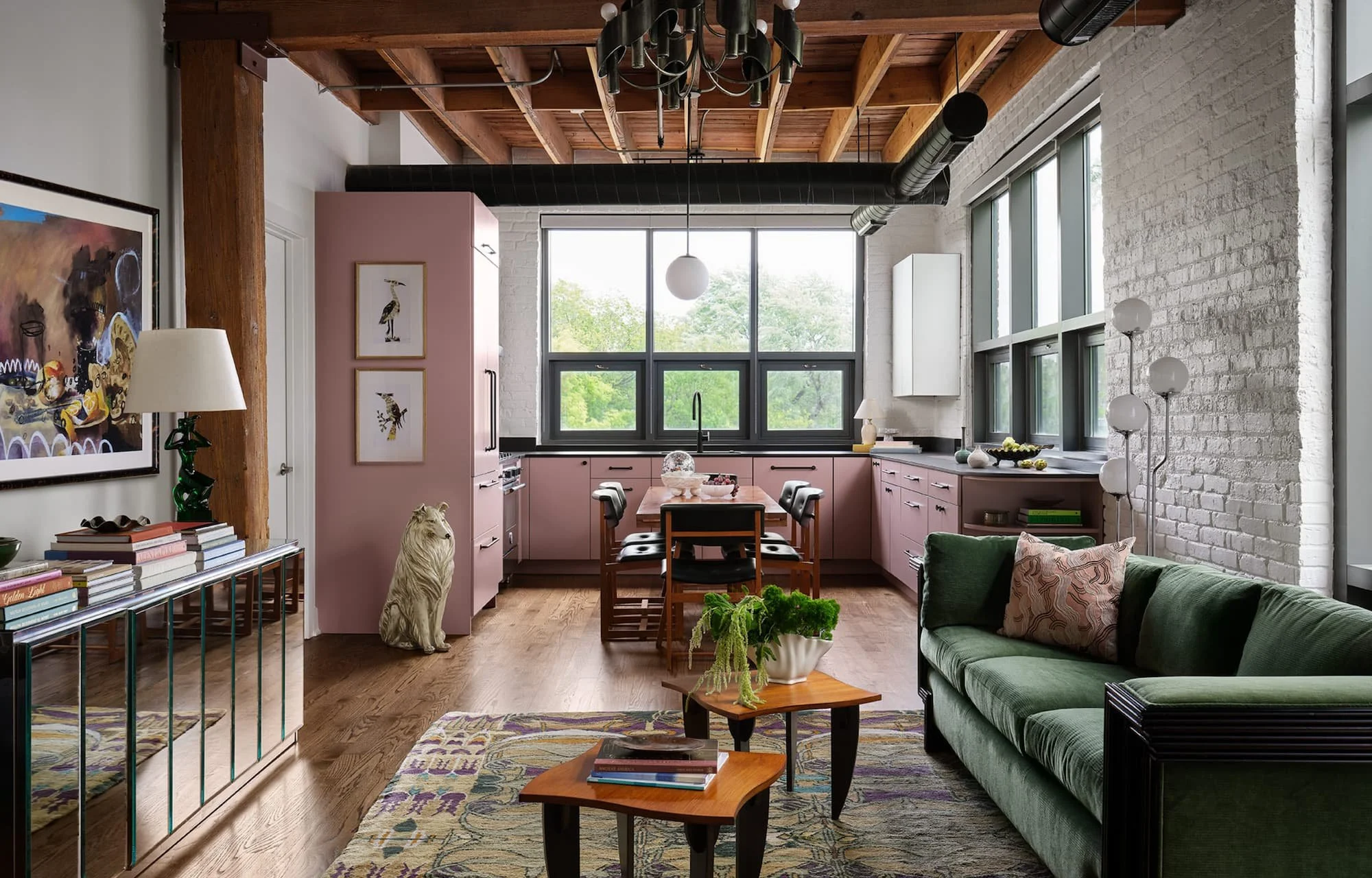 Modern living room with brick walls and large windows, featuring a green sofa, a pink kitchen area, artwork, and a sculpture of a lion near a table with potted plants.