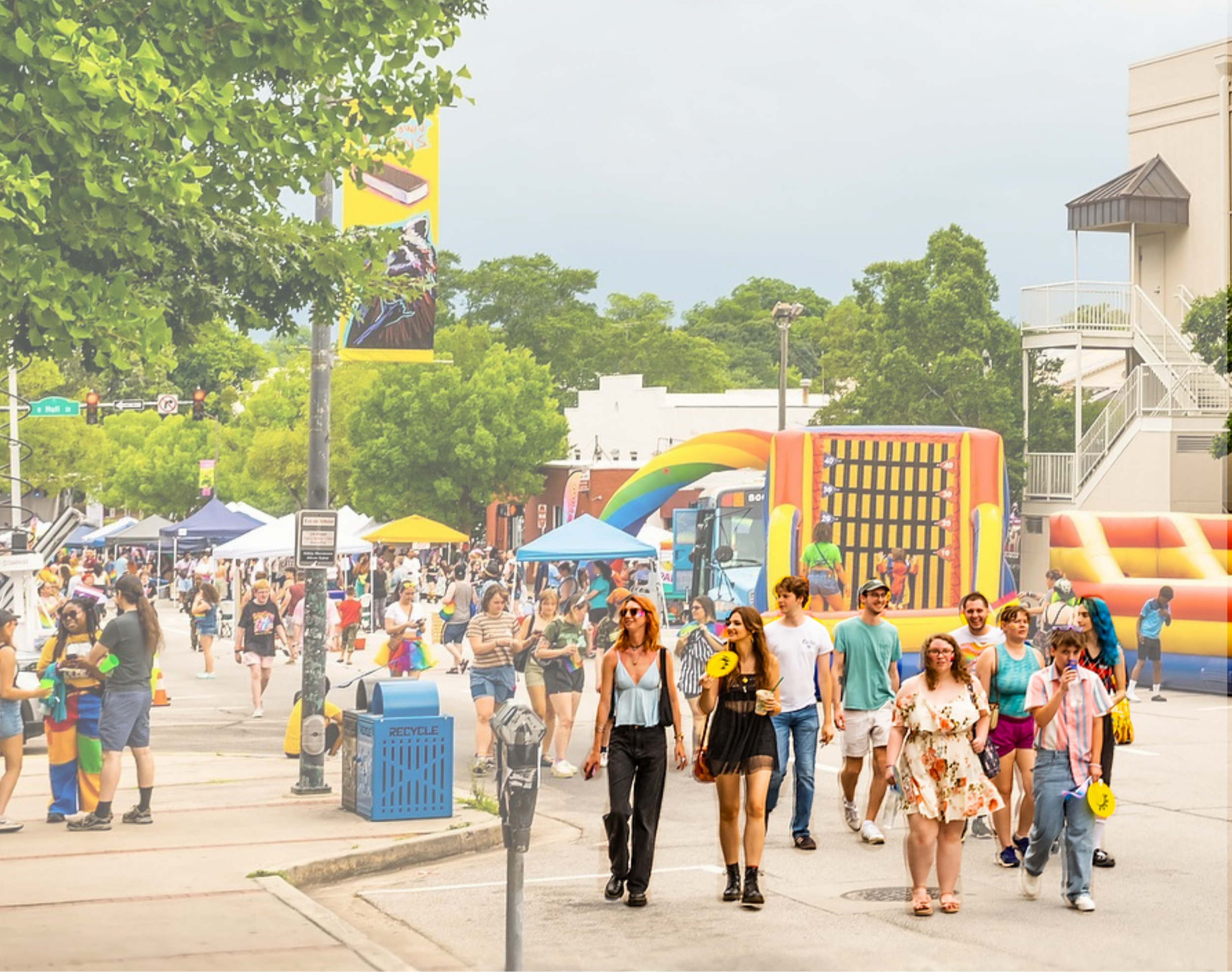 Crowd of people walking outdoors at PRIDE, with tents, inflatable bounce houses, and inflatable rainbow arch visible in the background.