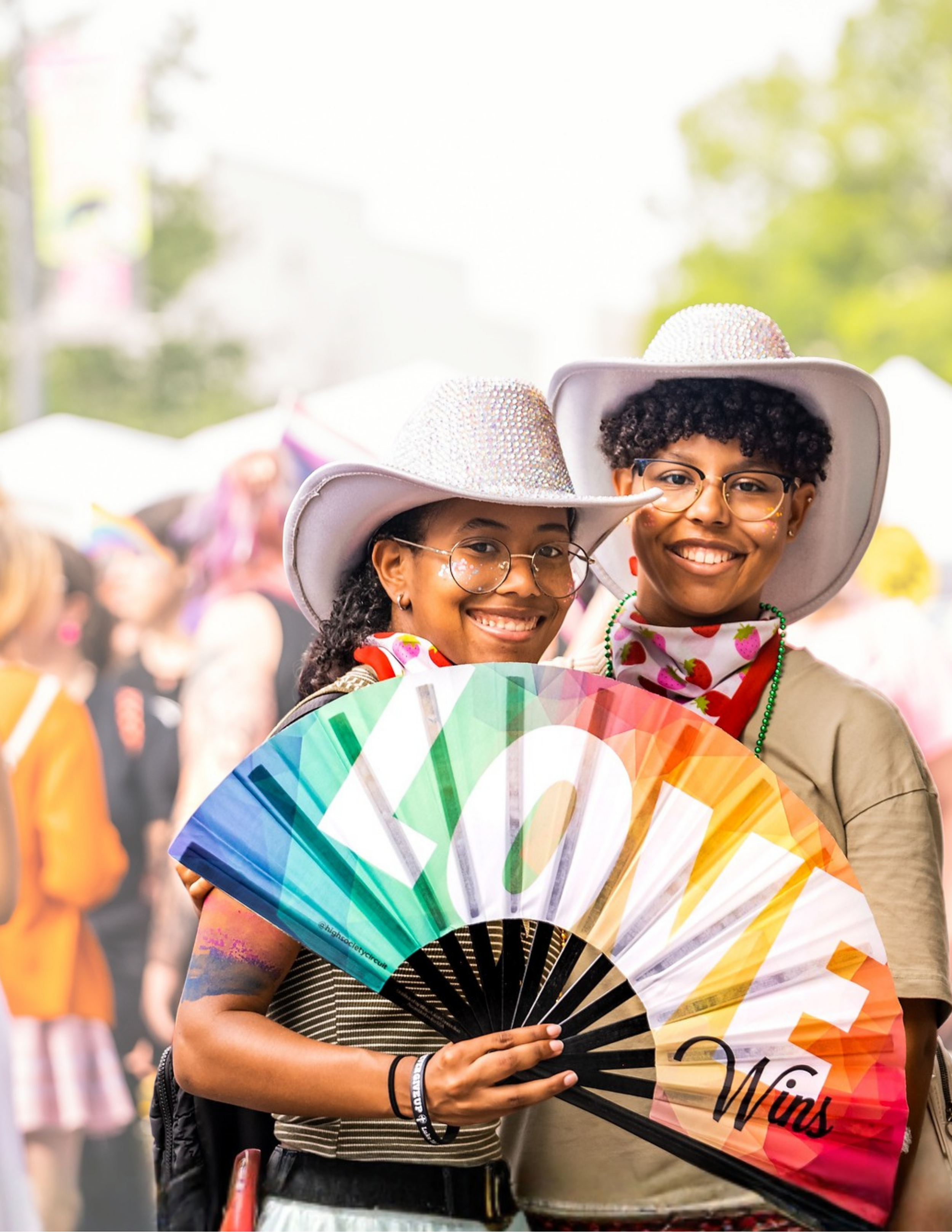 Two smiling people wearing white cowboy hats and glasses, holding a rainbow-colored fan with the words 'LOVE WINS' on it at PRIDE.