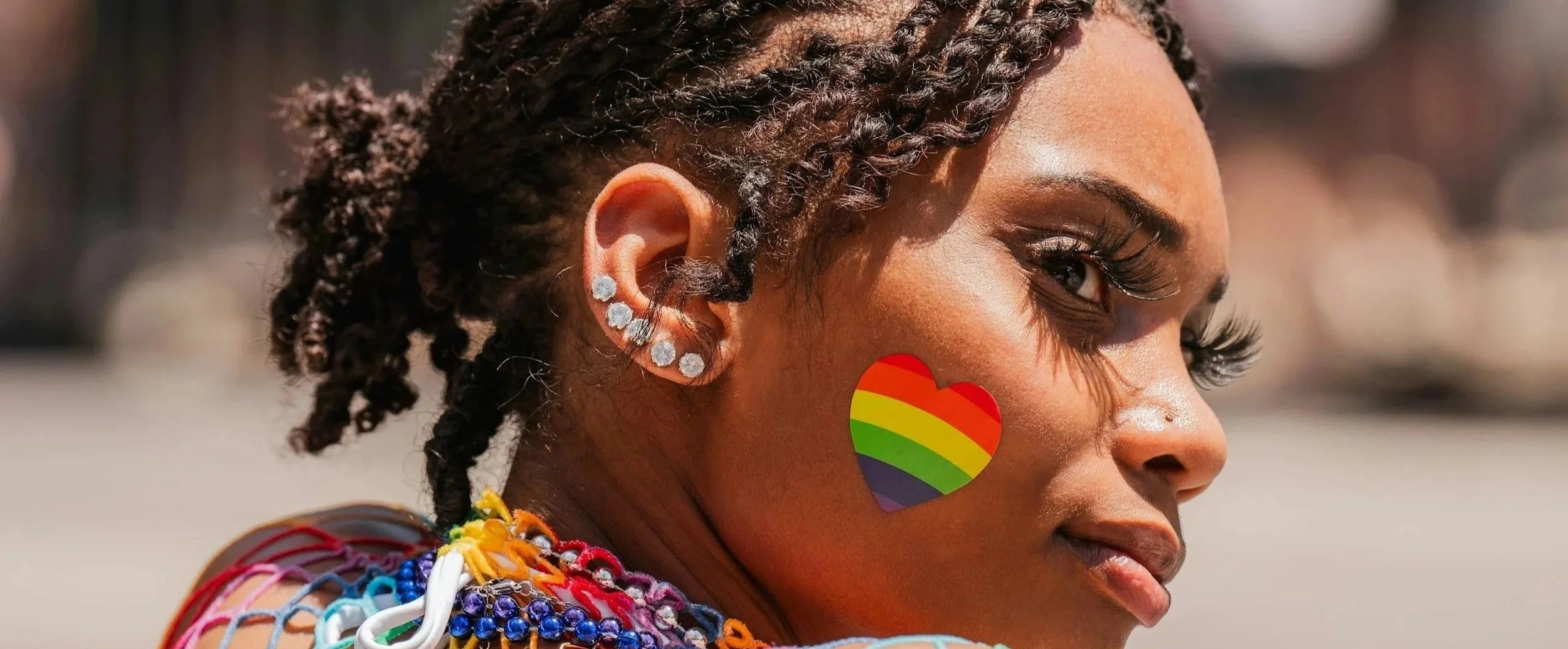 A person with biracial features, wearing multiple earrings with shiny stones, has colorful beads around their neck, a rainbow heart sticker on their cheek, and wears makeup with long eyelashes.