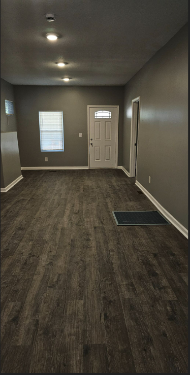 Empty living room with gray walls, dark wood flooring, two windows, a front door, and a floor vent.