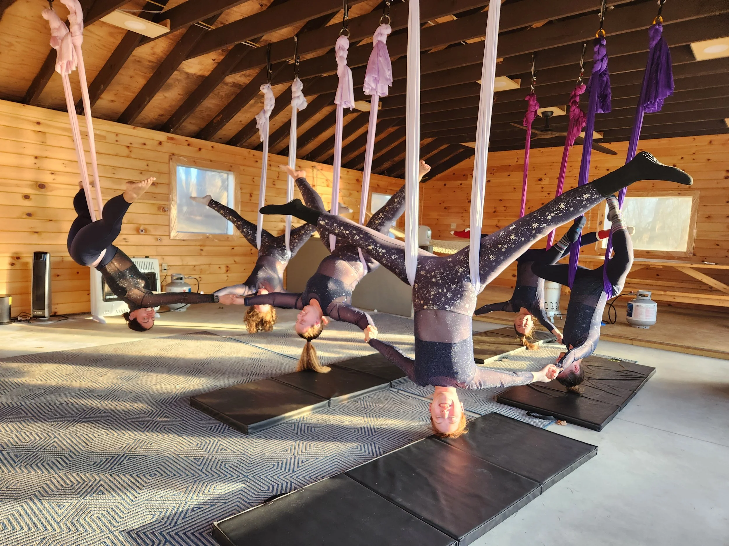 Five women in matching black and star-patterned athletic wear hanging upside down on aerial silk hammocks in a wooden studio, holding hands and smiling.