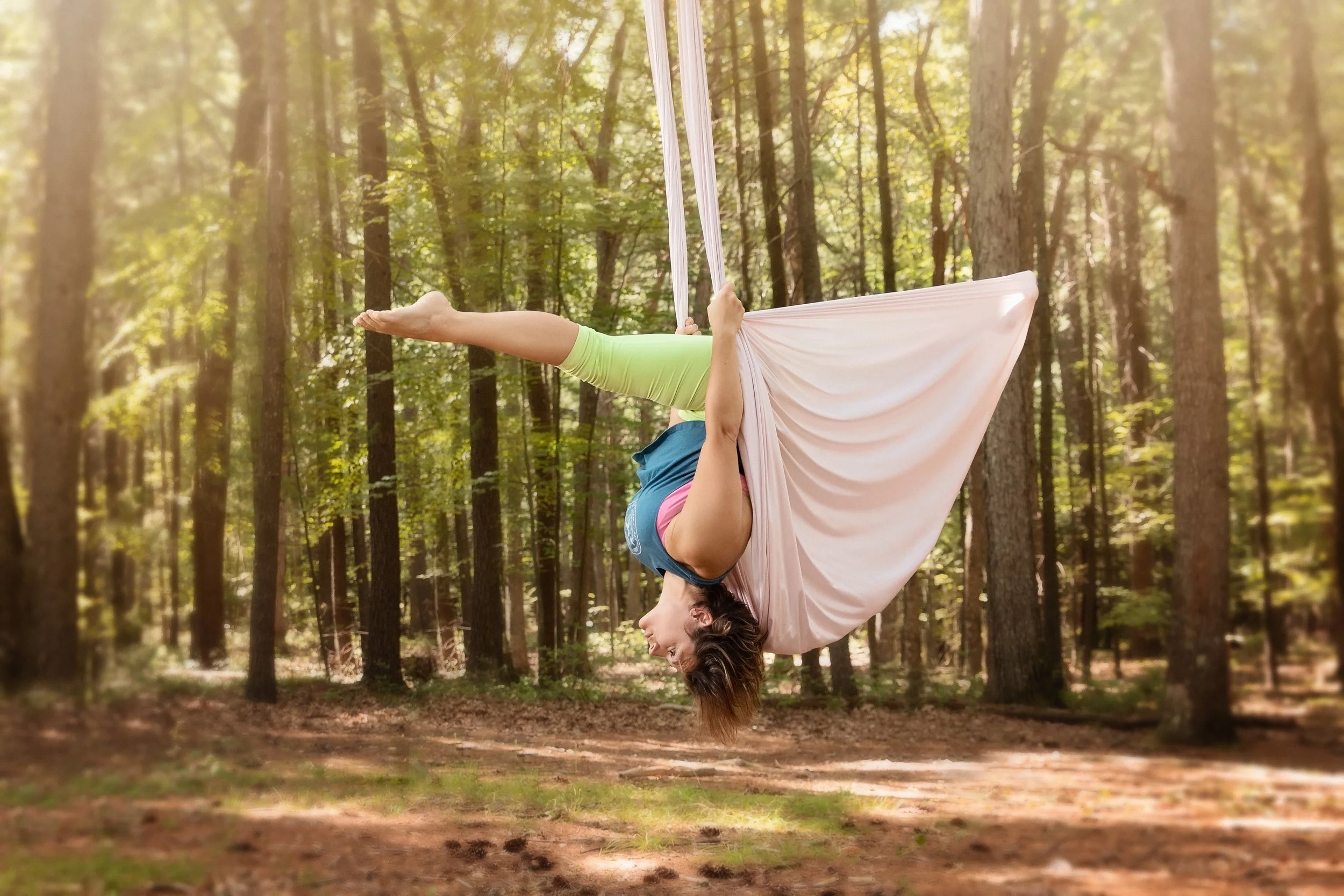 A woman practicing aerial silk yoga in a forest, upside down with her head near the ground, hanging from pink fabric.