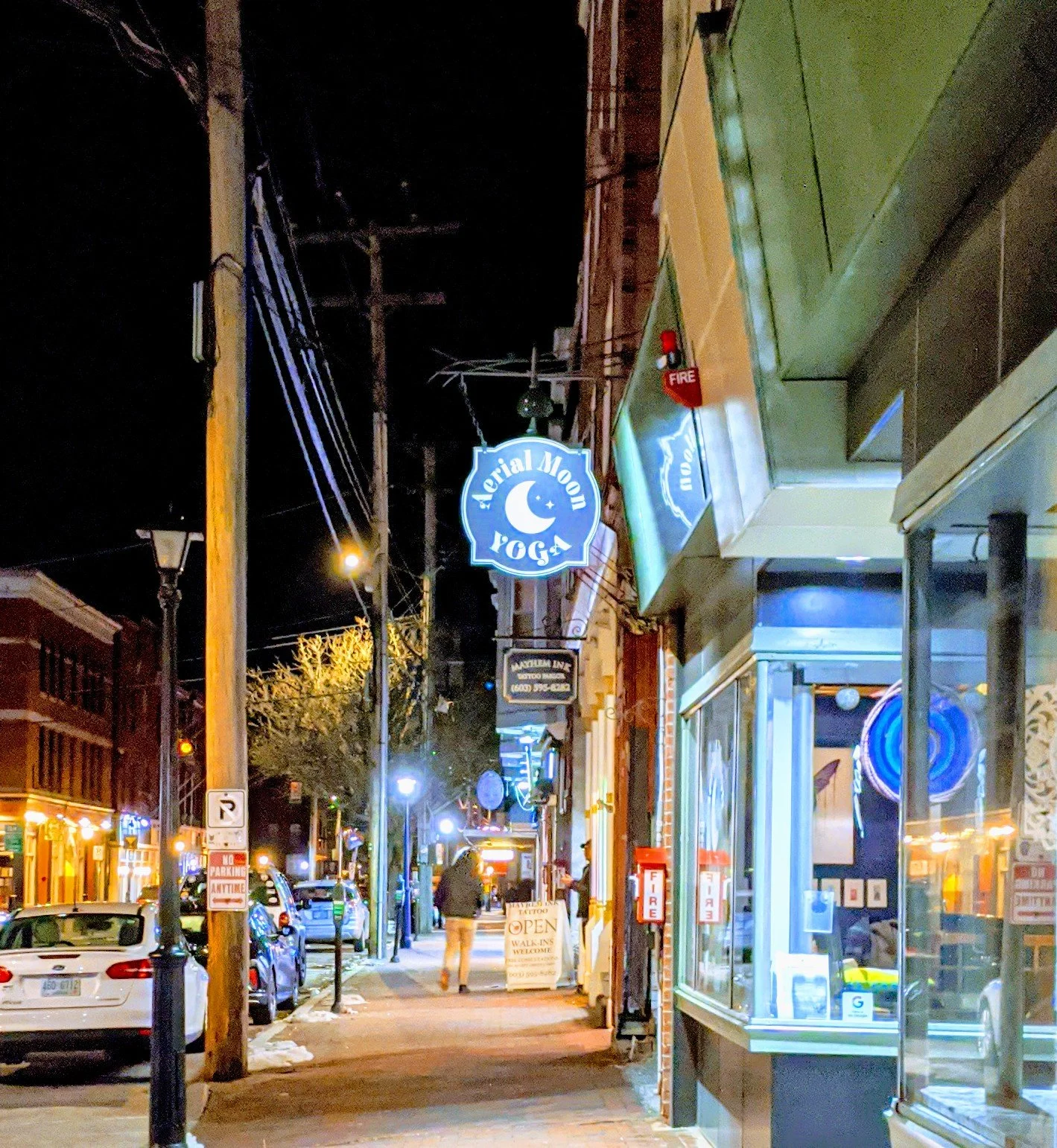 Nighttime street scene with parked cars, illuminated storefronts, and signs including a yoga studio sign and a tattoo shop sign. Pedestrians walk along the sidewalk.
