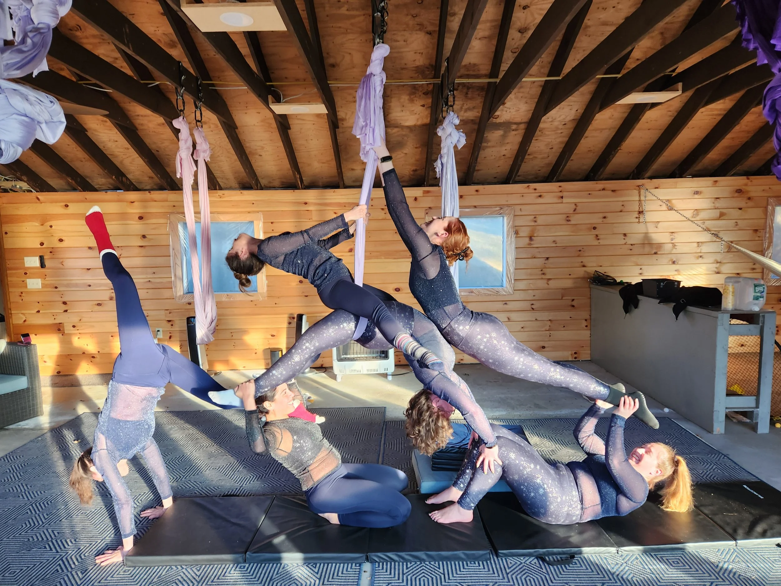 A group of women practicing acro yoga indoors, with one woman on the floor supporting two women balancing above her, who are holding onto fabric hammocks attached to the ceiling.