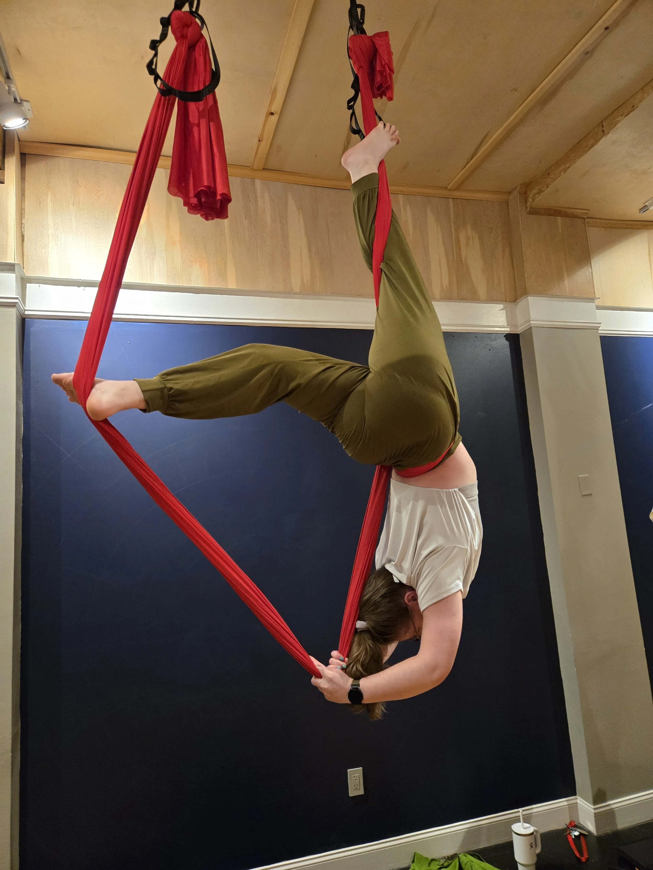 A woman performing aerial yoga on red fabric hammocks inside a room with wooden walls and ceiling.