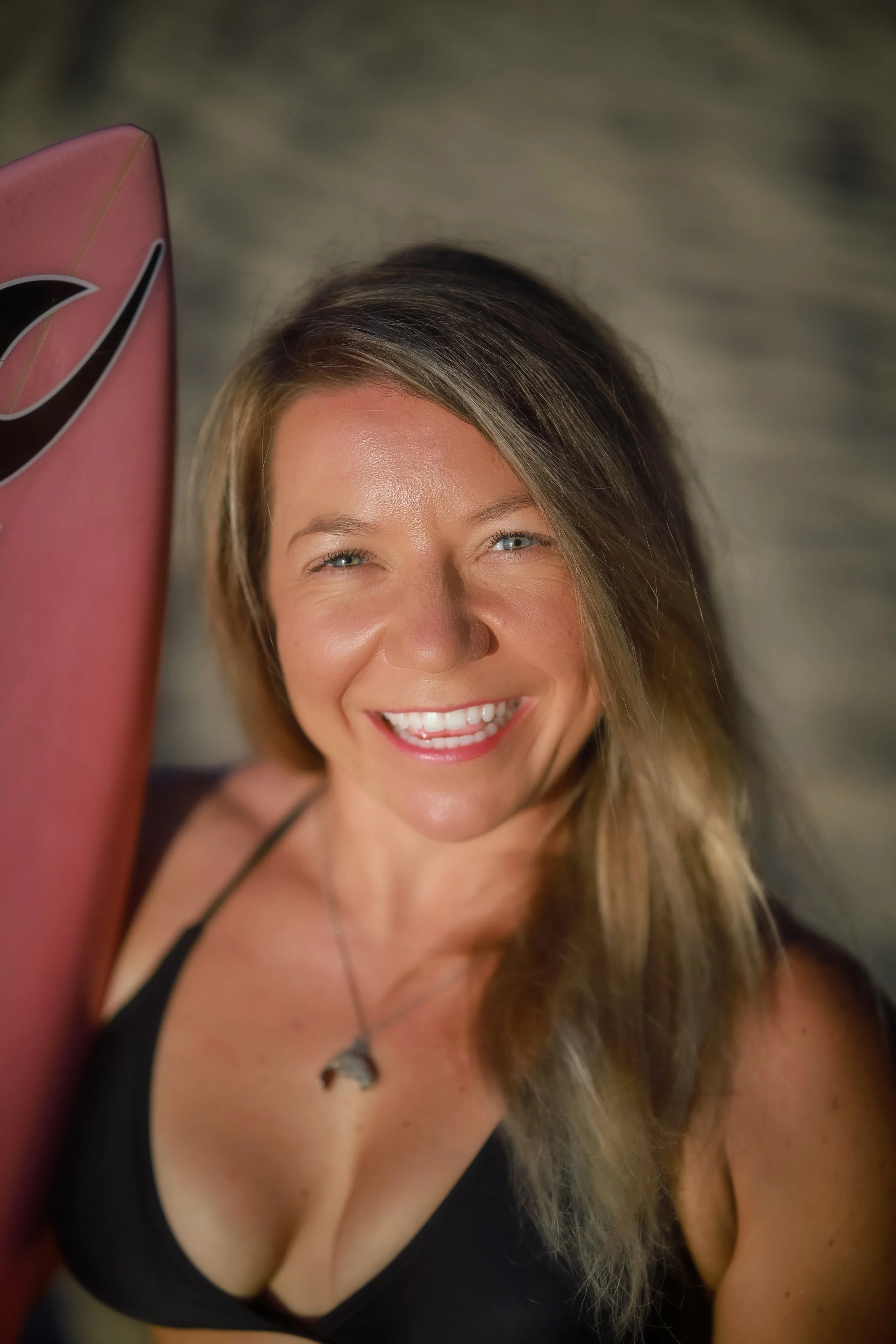 A smiling woman with blonde hair and blue eyes wearing a black top and a necklace, holding a pink surfboard.
