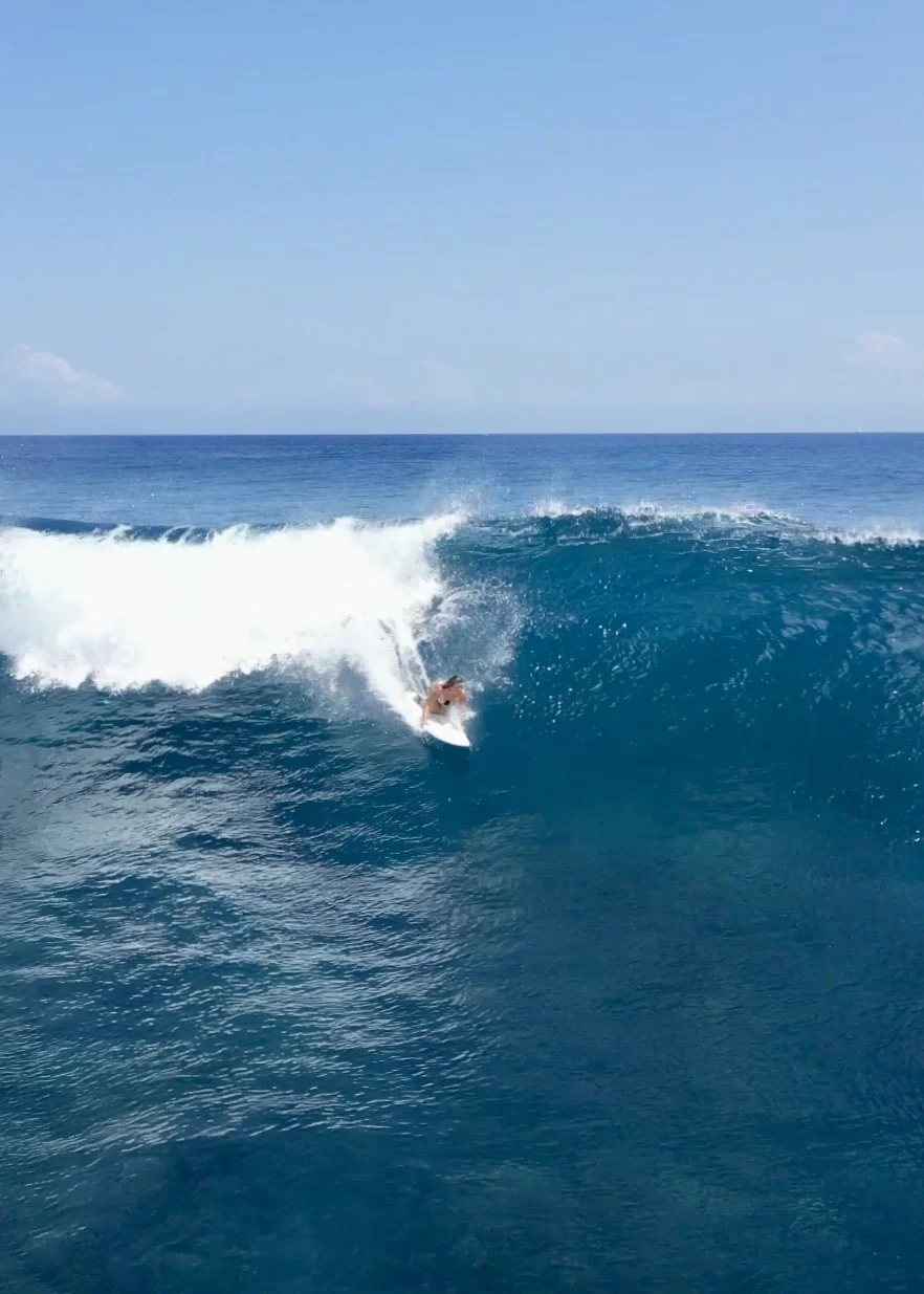 A person surfing on a large blue wave in the ocean under a clear sky.