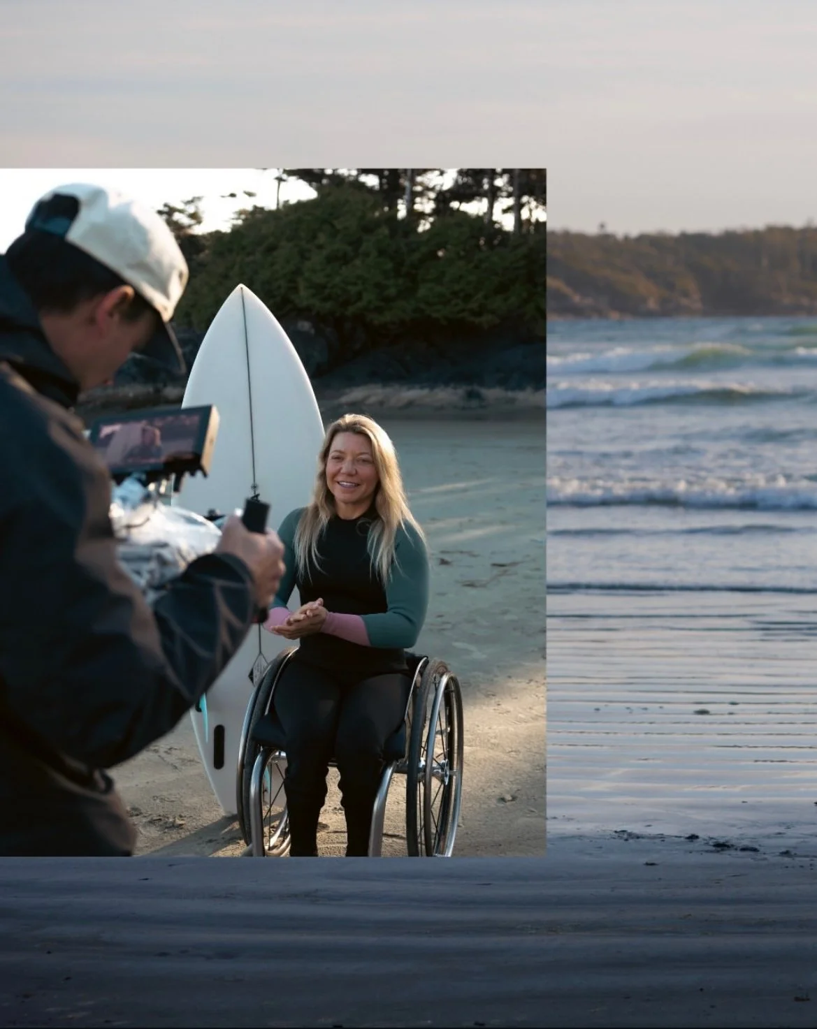 Person in a wheelchair being filmed or photographed on a beach with a paddleboard and ocean in the background.