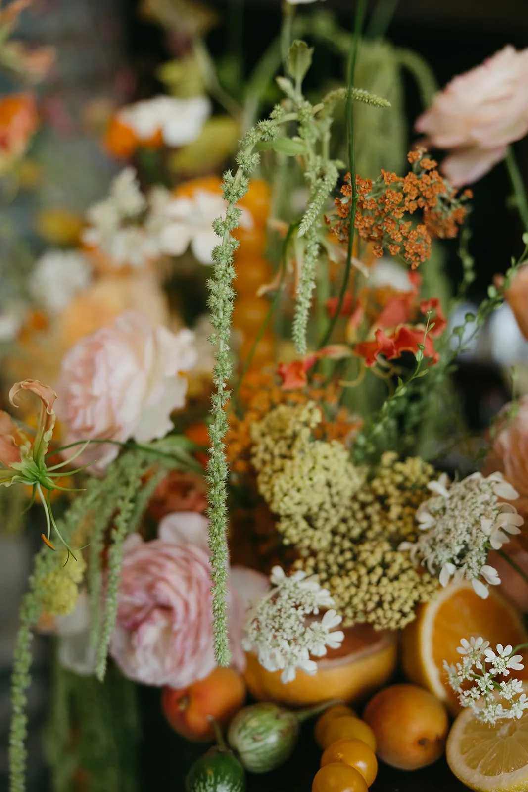 A vibrant, whimsical floral arrangement with pink garden roses, orange blooms, and green amaranthus accented by halved citrus fruits and small kumquats.