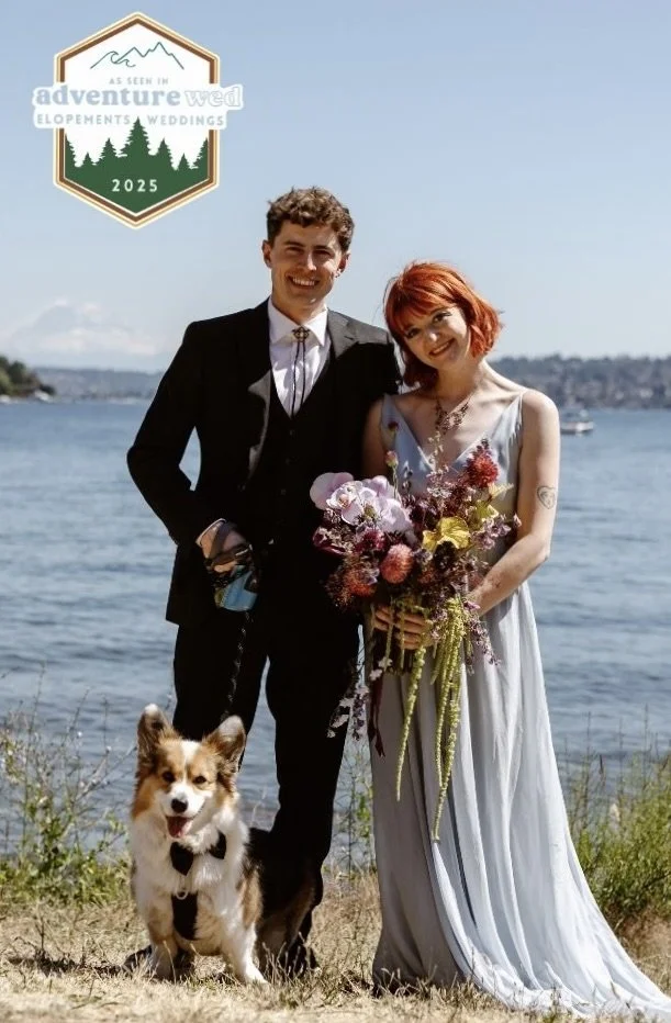 A couple who just eloped standing in front of the Puget Sound water with their corgi puppy. Bride is wearing a light blue dress and holding a whimsical bouquet designed by New Leaf Floral Design.
