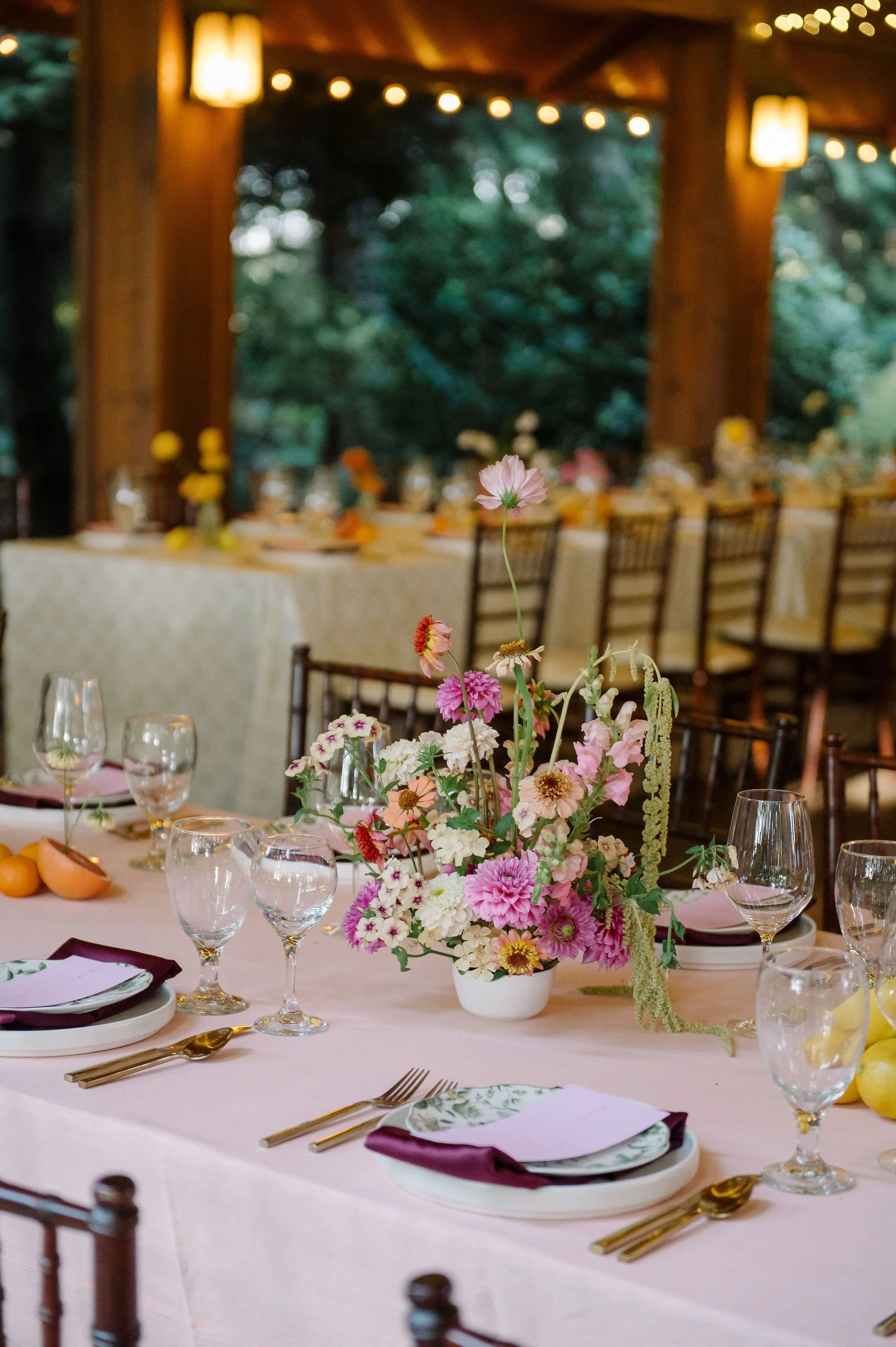 A lush ikebana inspired centerpiece at a wedding reception at Evergreen Gardens in Bellingham, featuring local dahlias and snapdragons in a palette of peach, blush, pinks, and chartreuse green on a long table with a blush pink table cloth.