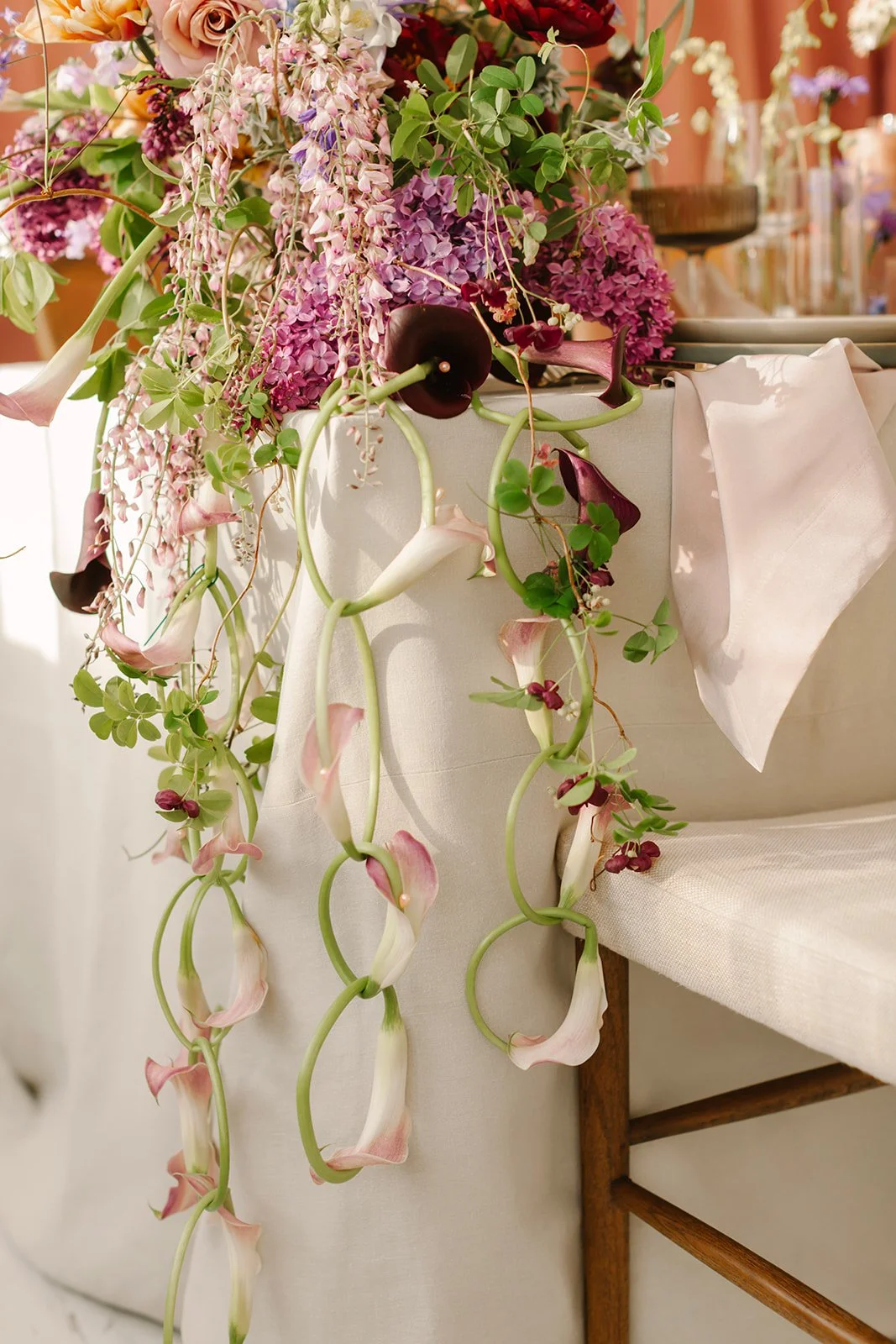 Cascading lavender and plum calla lily chains spill elegantly over the side of an ivory clothed table from a lush centerpiece full of spring flowers such as wisteria and tulips in a color palette of deep purples, vibrant lavender and soft mauves.