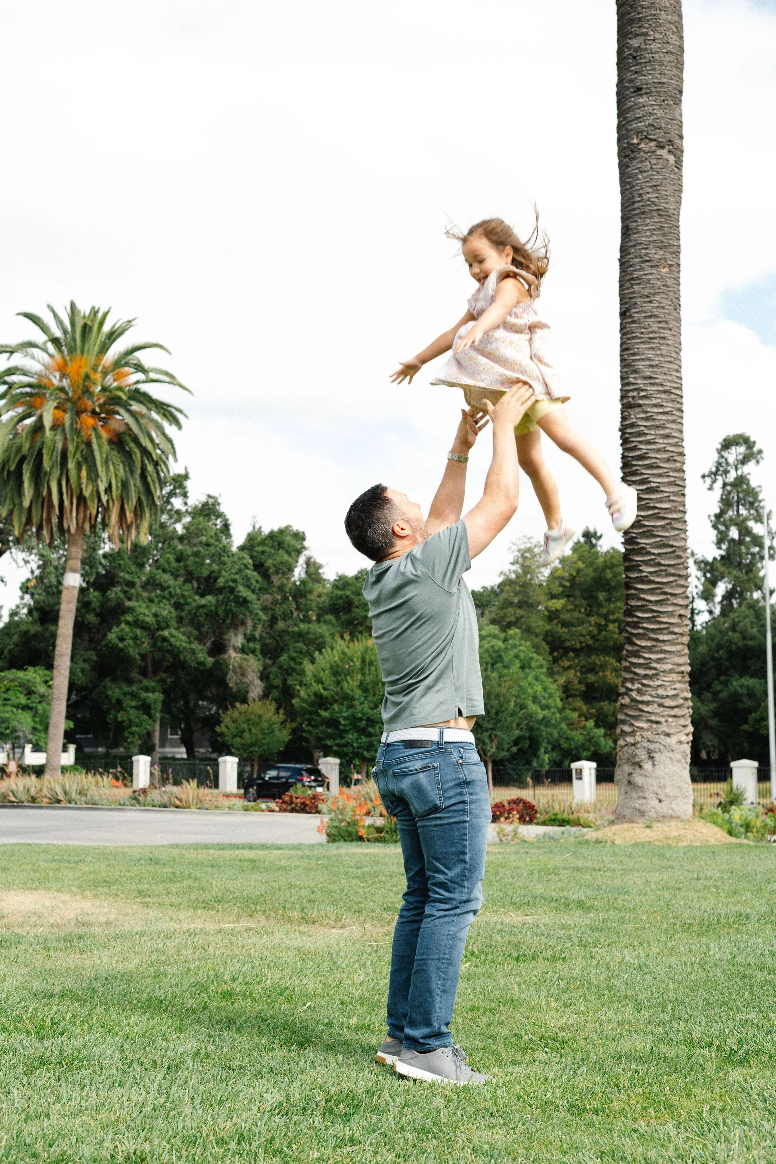 A man lifting a young girl into the air outdoors in a grassy park with palm trees and other greenery in the background.