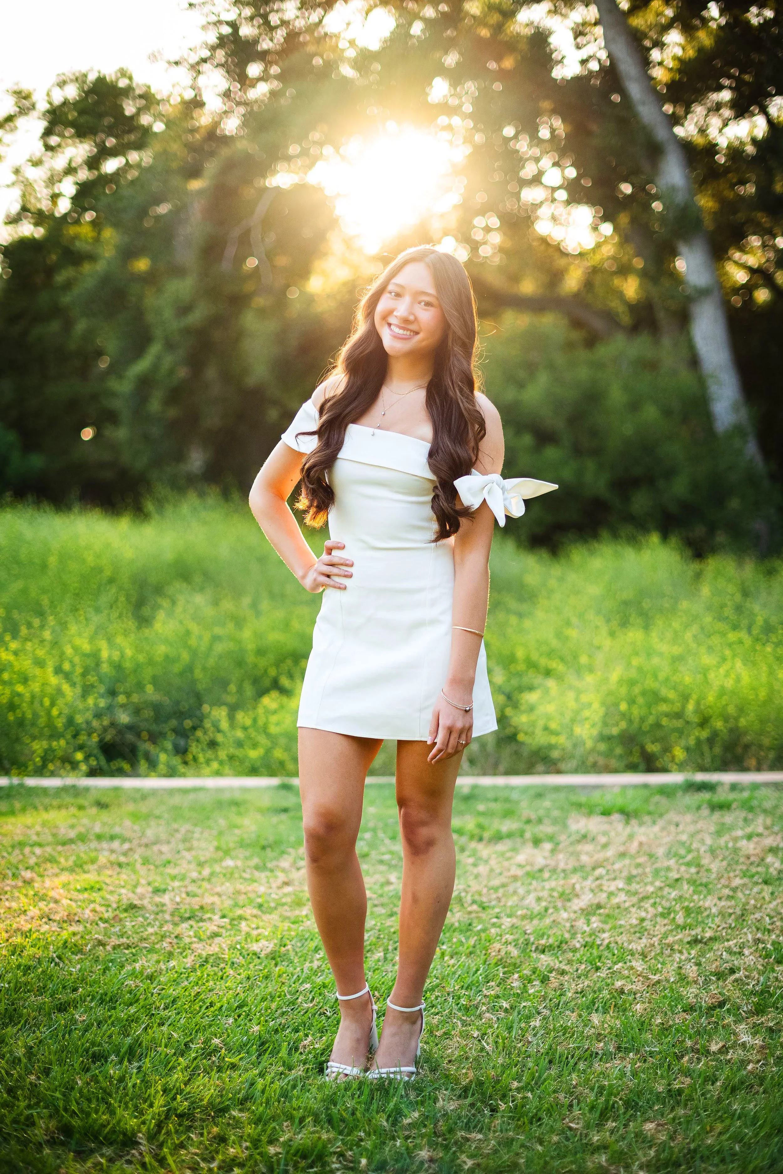 A young woman with long dark hair, smiling, wearing an off-shoulder white dress with a bow on the sleeve, standing outdoors on a grassy area with green trees in the background, backlit by the setting sun.