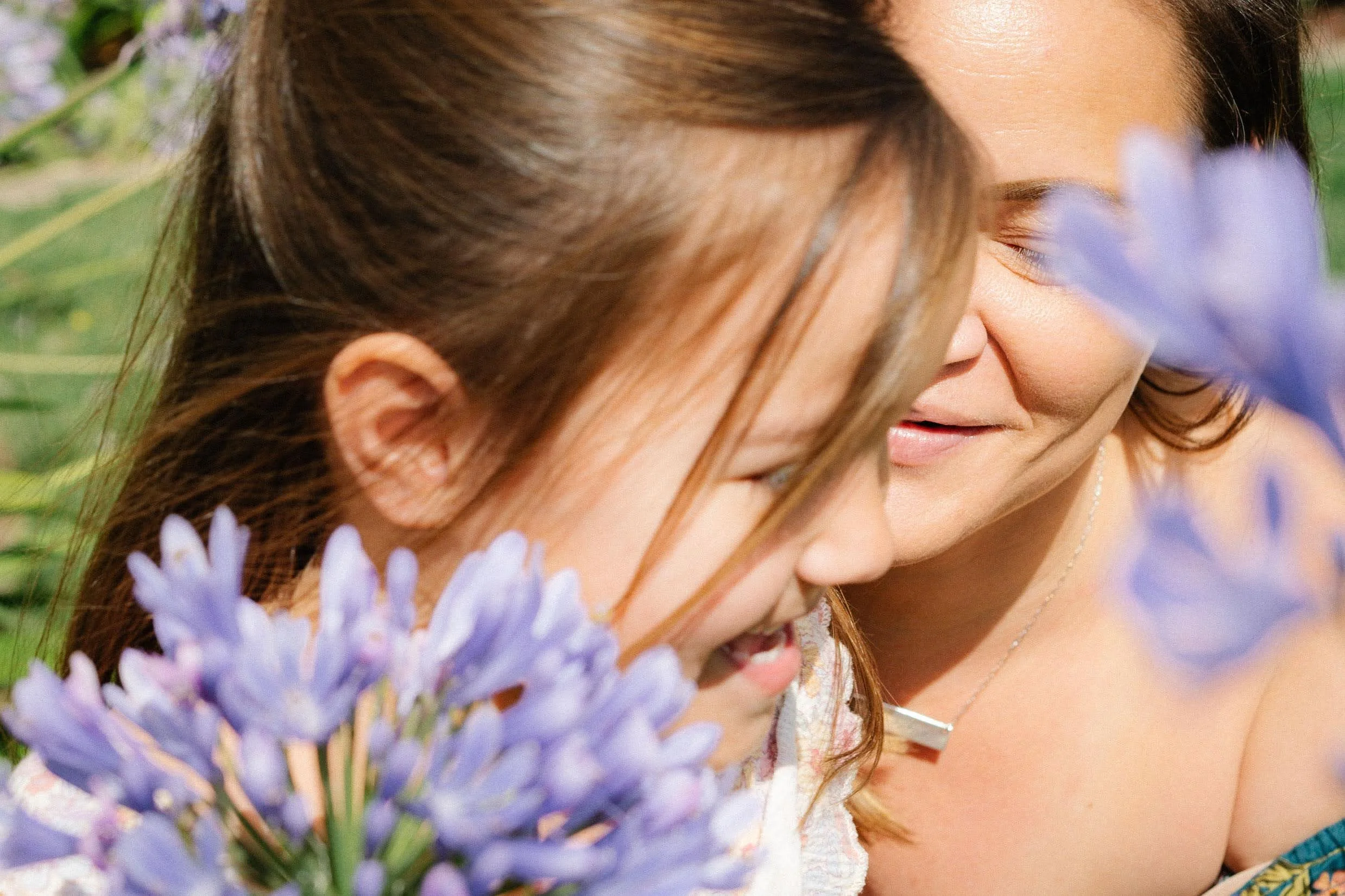 Close-up of a woman and a girl sharing a moment among purple flowers, both smiling and with eyes closed.