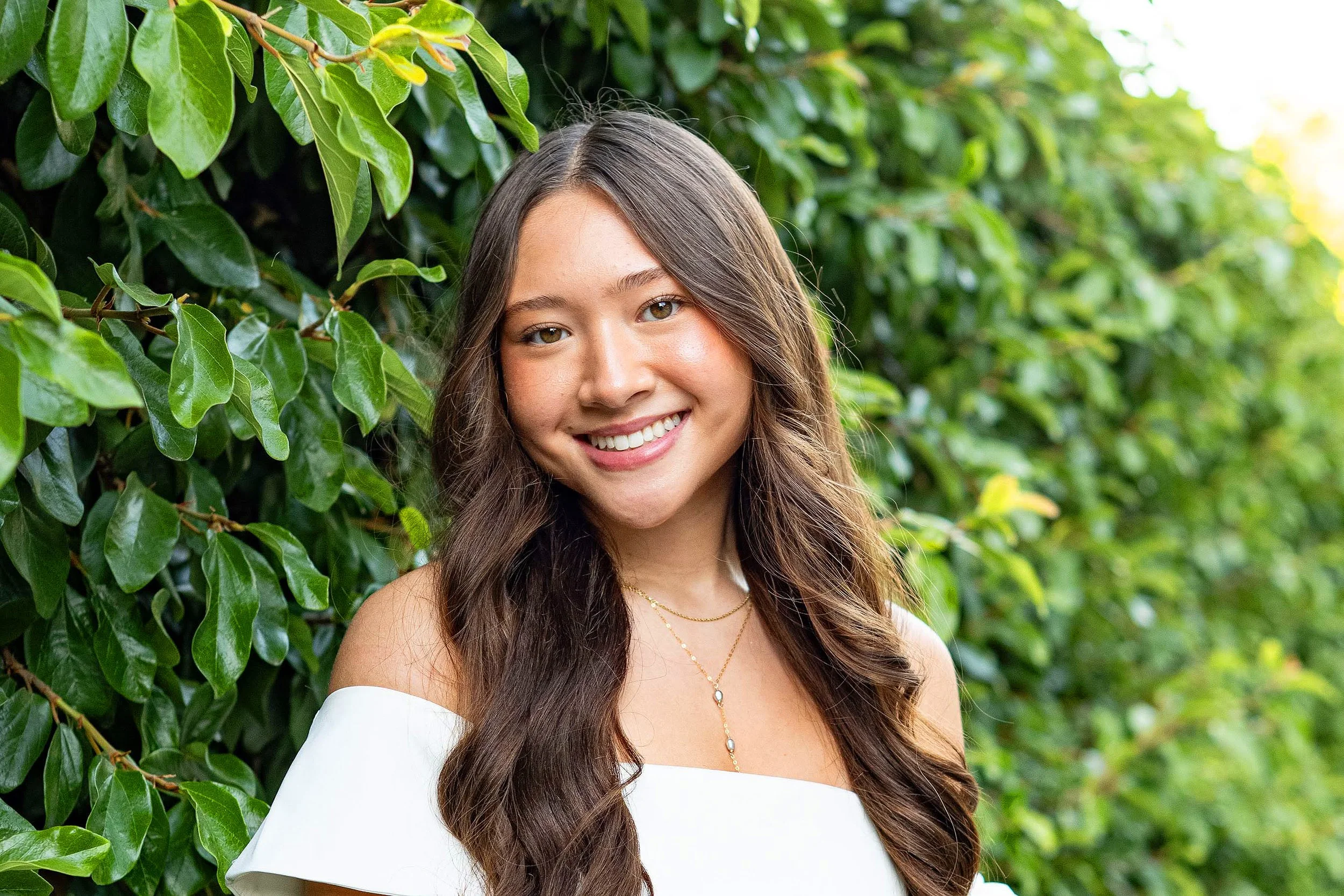 A young woman with long brown hair and tan eyes, smiling, standing in front of green bushes with shiny leaves.
