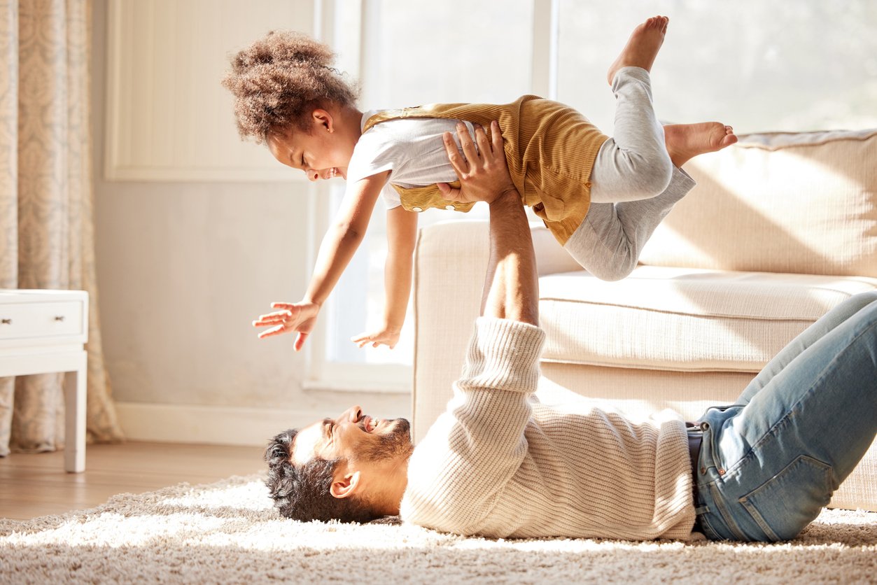 Father laying with back on the floor holding his young daughter as if she is flying. They appear joyful.