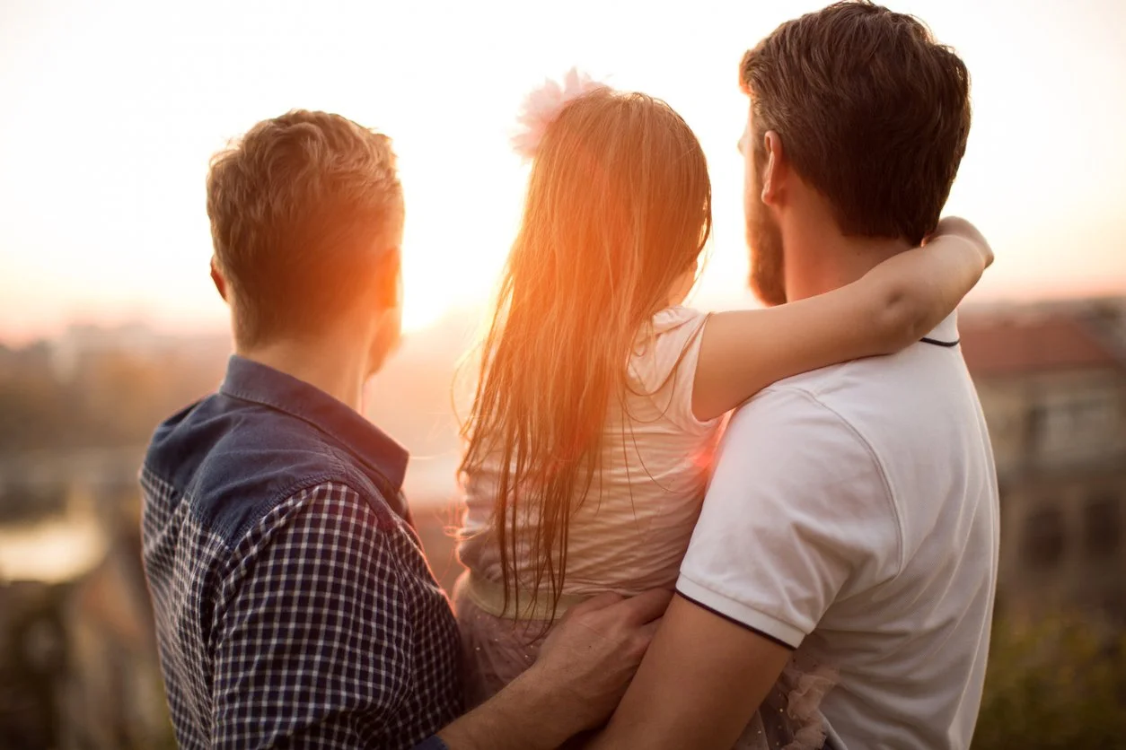 Two dads holding their daughter and looking peacefully at a sunset.