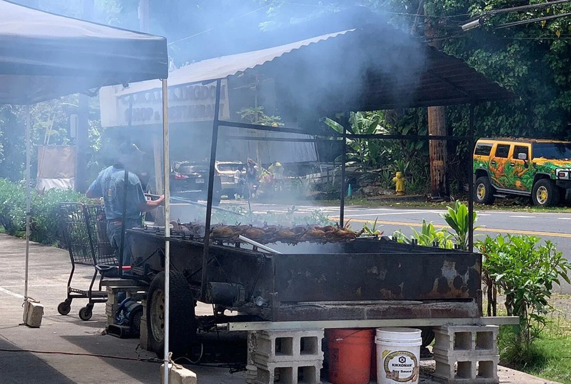 Street food vendor cooking meat on a grill under a canopy with cars parked in the background and a decorated vehicle on the roadside.