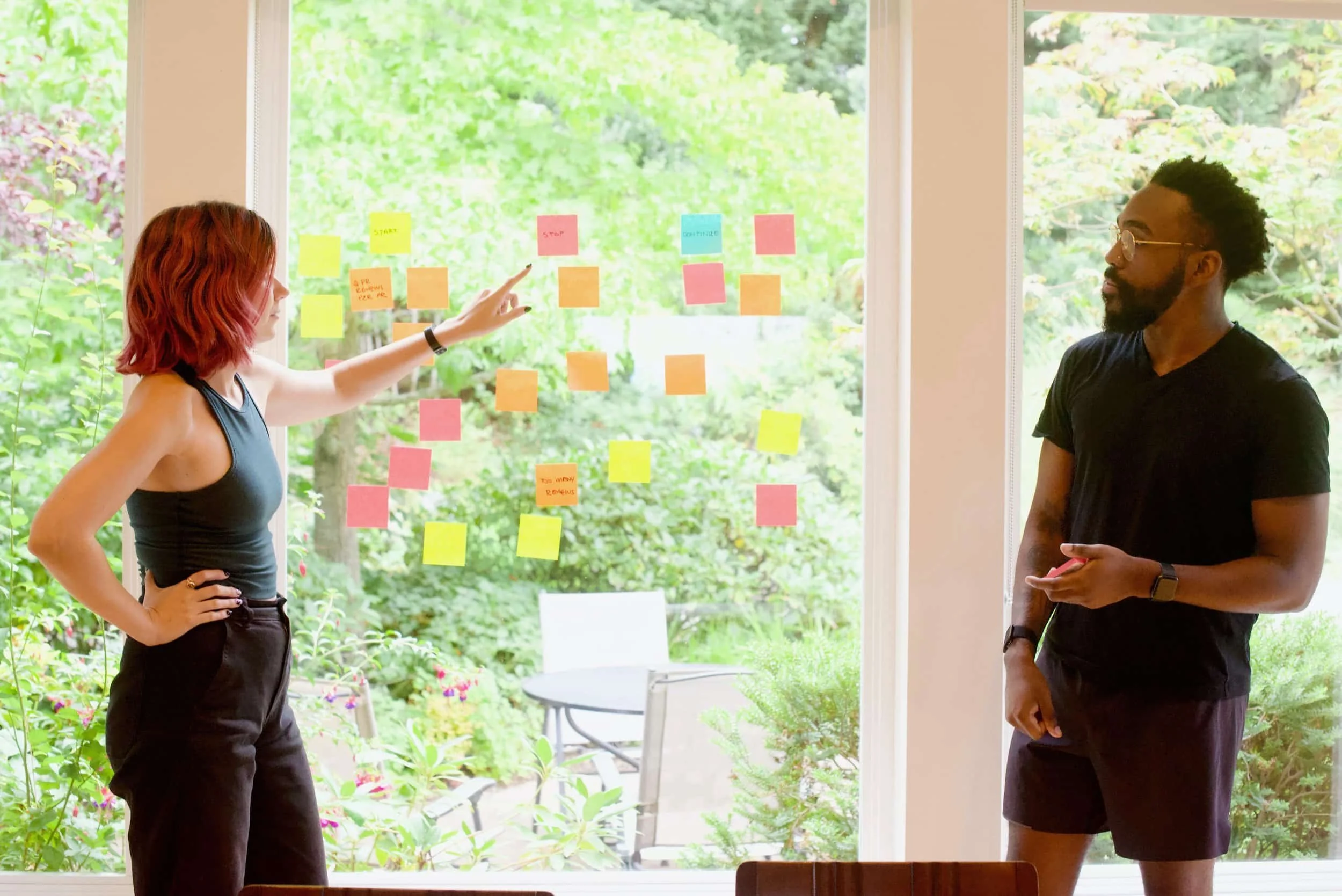Two people discuss colorful sticky notes on a large window, brainstorming green funding ideas with a lush garden visible outside.