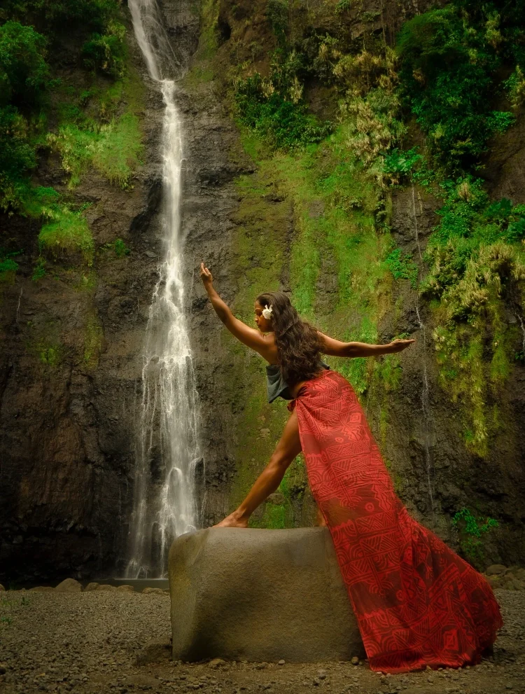 A Tahitian dancer in a red skirt and black top standing on a large rock in front of a waterfall, with her arms outstretched and one arm pointed upward, in a lush green natural setting.