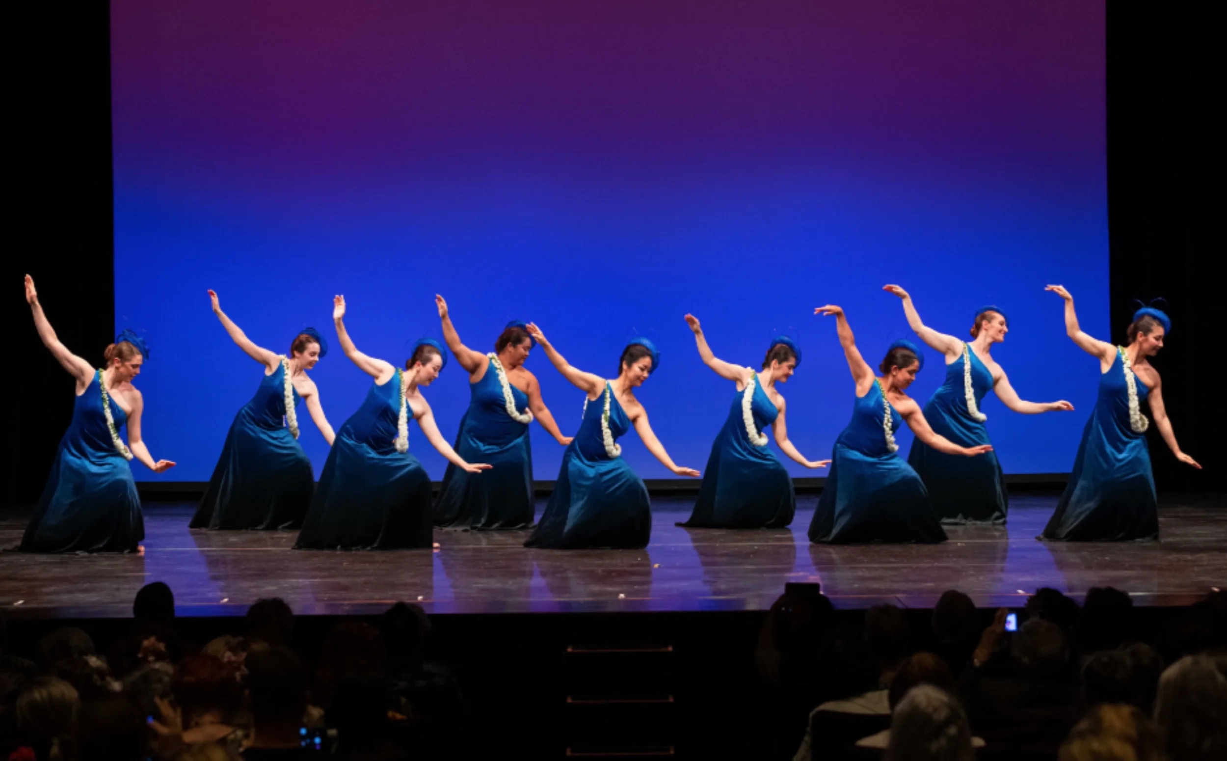 Ten women in blue dresses Hula dancing on stage with a blue background, wearing leis and blue hair accessories, performing a synchronized dance.