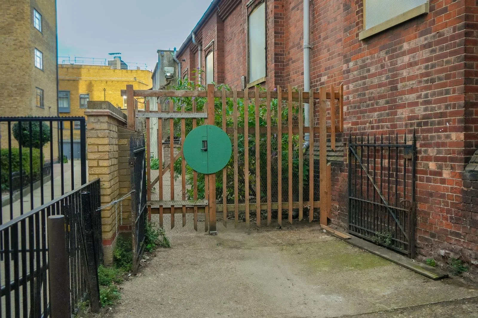 A small outdoor area enclosed by a wooden fence and brick walls, with a green round electrical or utility box mounted on the fence. There is a small metal gate on the right side, and the ground is dirt and concrete. Behind the fence, there are green plants and a narrow alleyway between buildings.