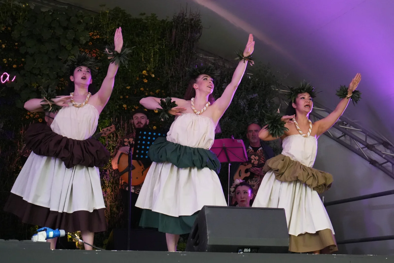 Three women wearing traditional hula skirts and leis performing a dance on stage with musicians in the background, decorated with greenery and flowers.