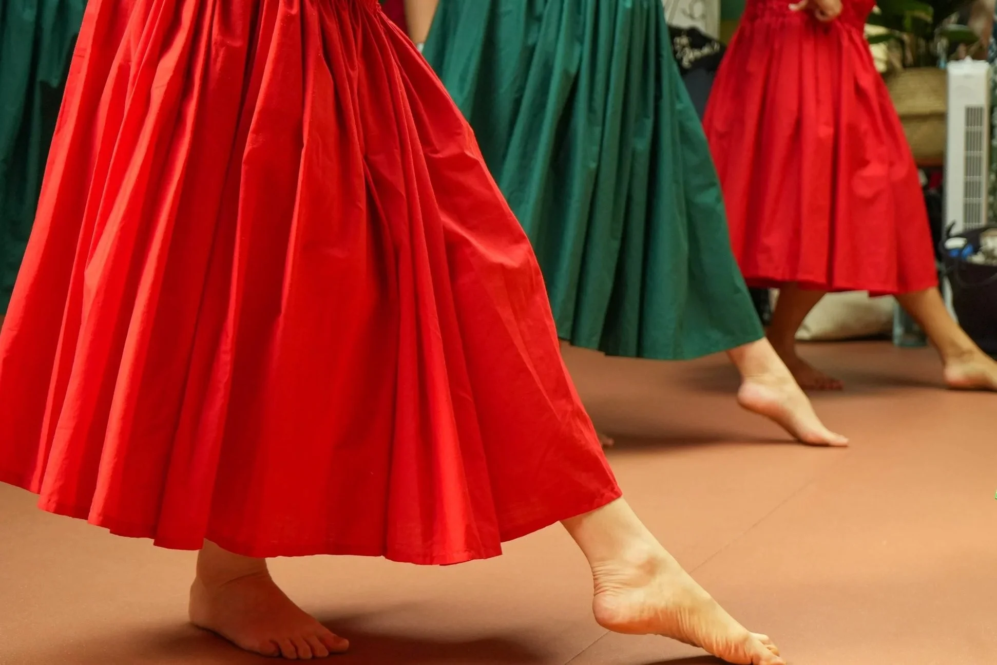 Close-up of Hula dancers in red and green skirts performing, focusing on their bare feet and flowing skirts.