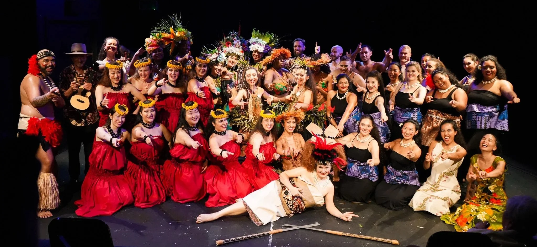 Group photo of performers dressed in traditional Polynesian costumes, some with floral crowns and leis, on stage after a cultural performance.
