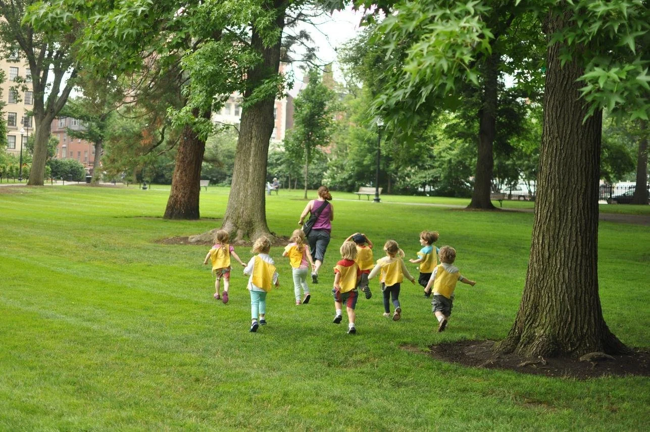 A group of young children wearing yellow vests running in a park with an adult supervising under a tree.