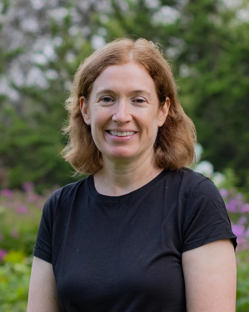 A woman with red hair, wearing a black T-shirt, smiling outdoors with green foliage and purple flowers in the background.
