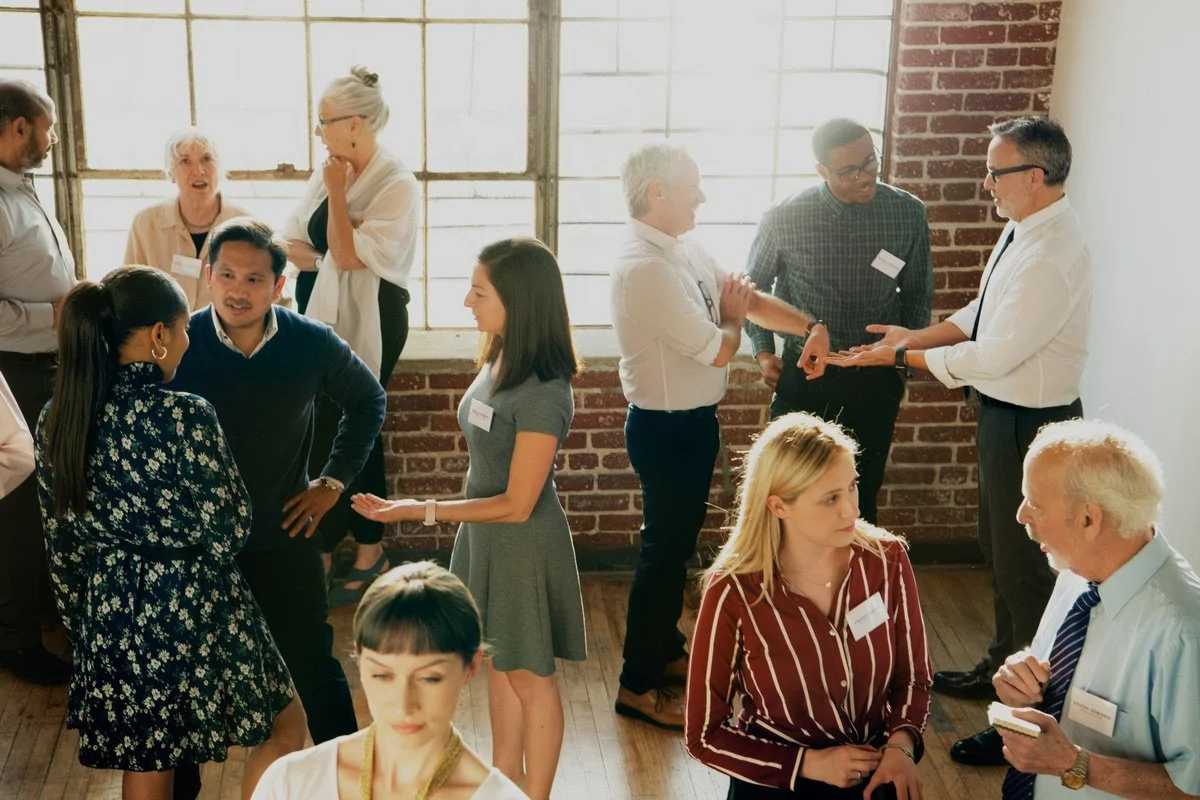 Group of people engaged in conversations at a professional networking event or conference in a room with brick and glass walls.