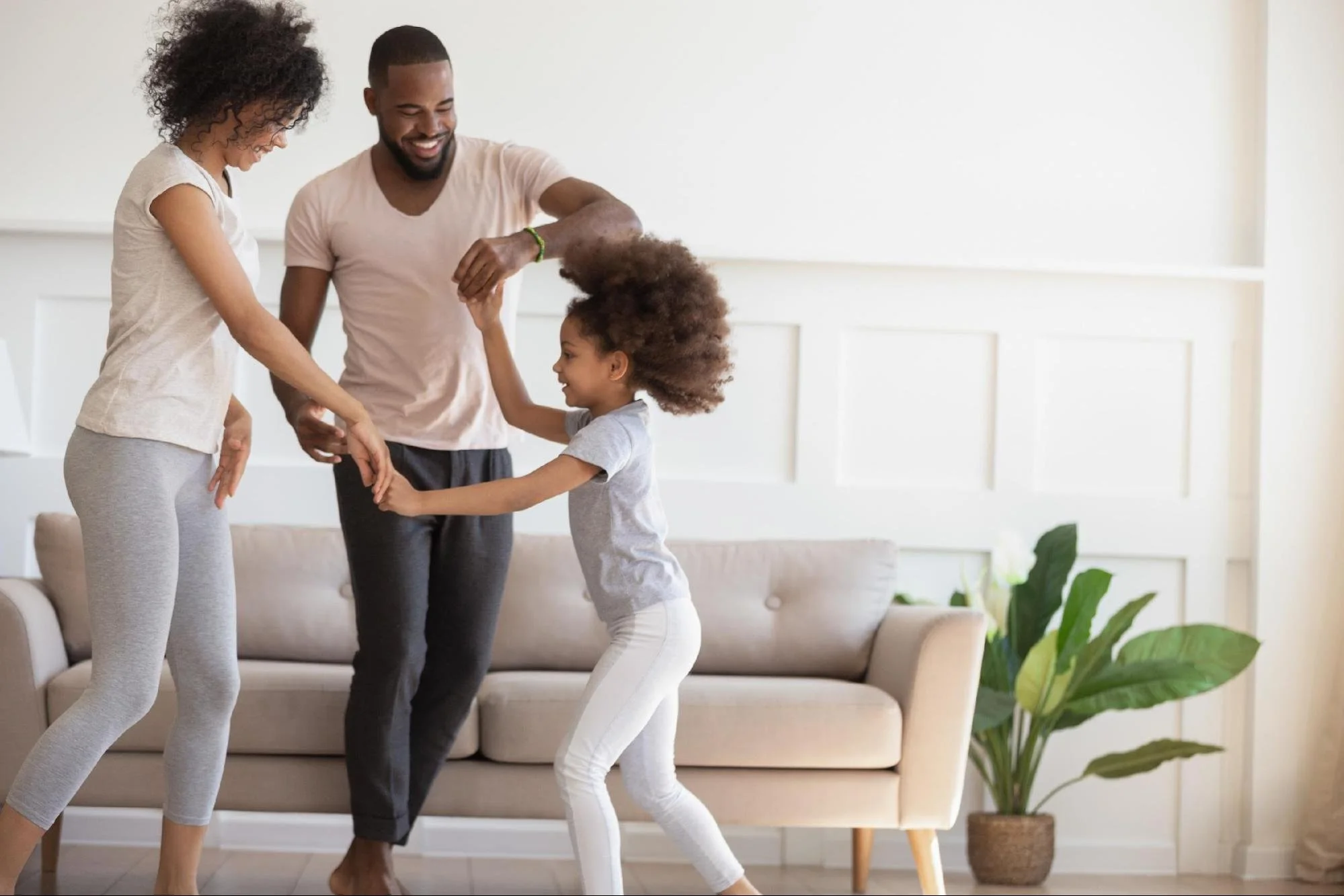 A happy family of three, a woman, a man, and a young girl, dancing and playing together in a bright living room with a beige couch and green houseplant.