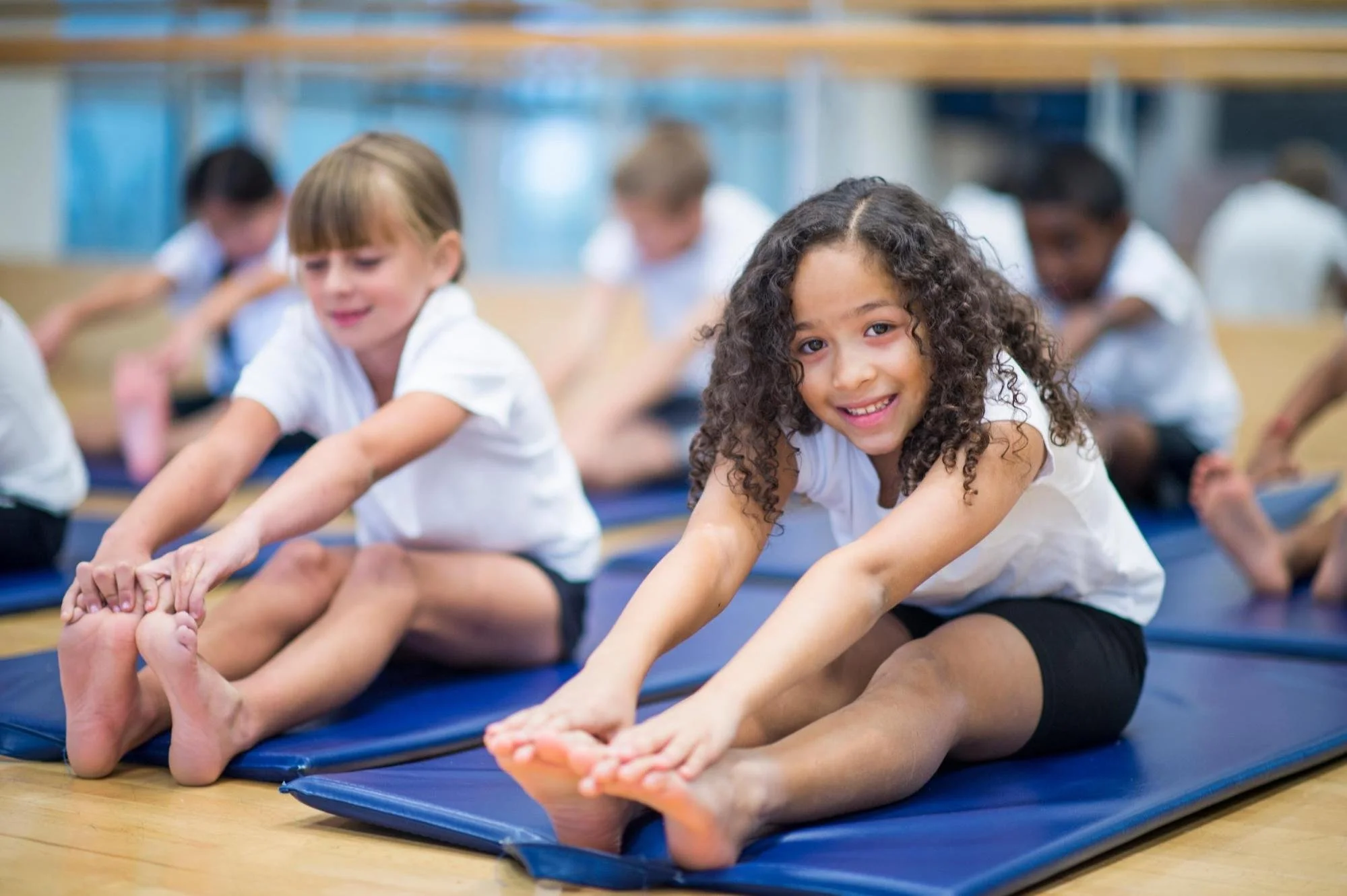 Group of children in white t-shirts and black shorts stretching on mats in a gym class.