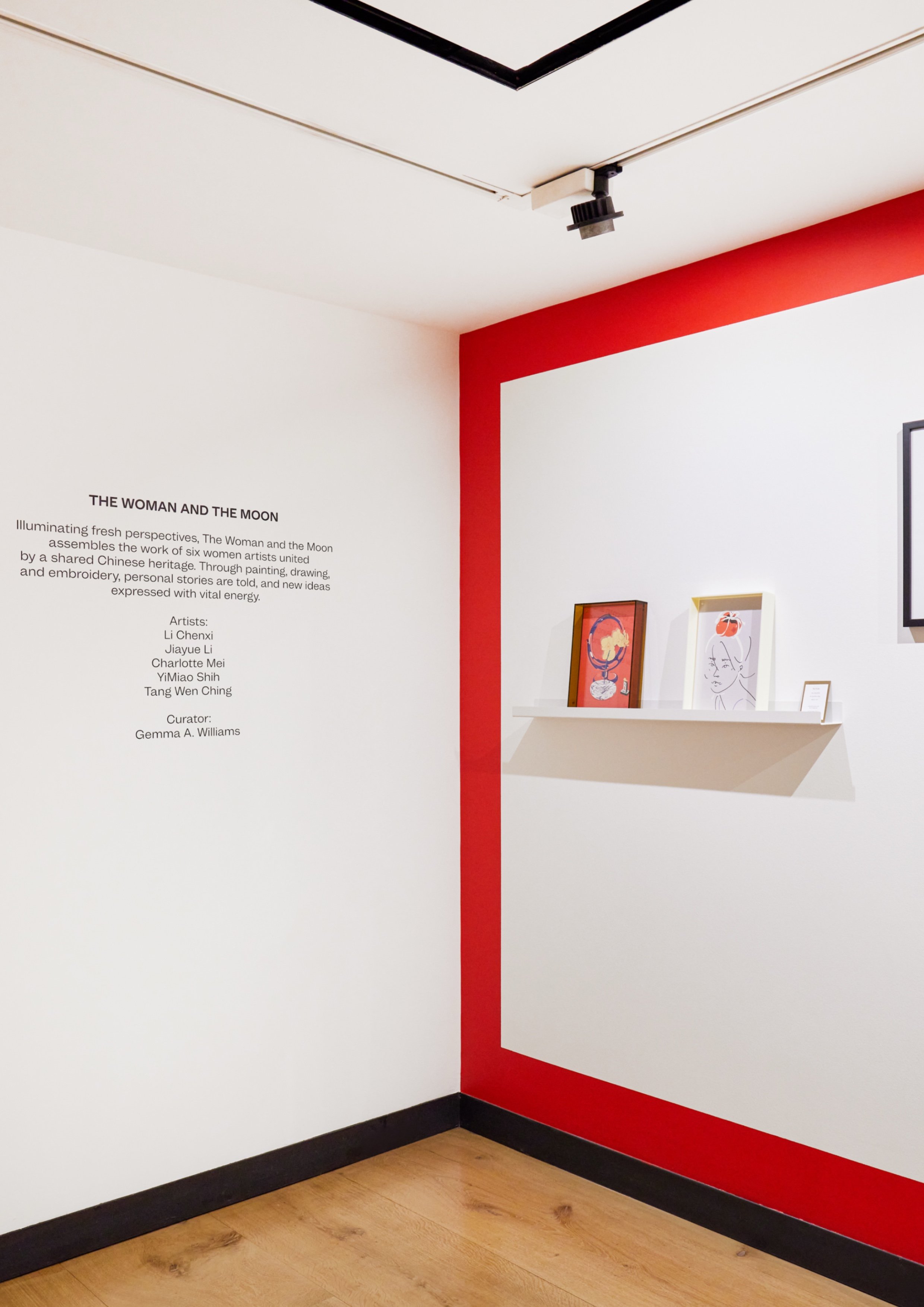 Interior photo of a white art gallery with a bold red painted rectangular frame on the wall, a floating shelf displaying two small artworks, and wall text reading "The Woman and the Moon"