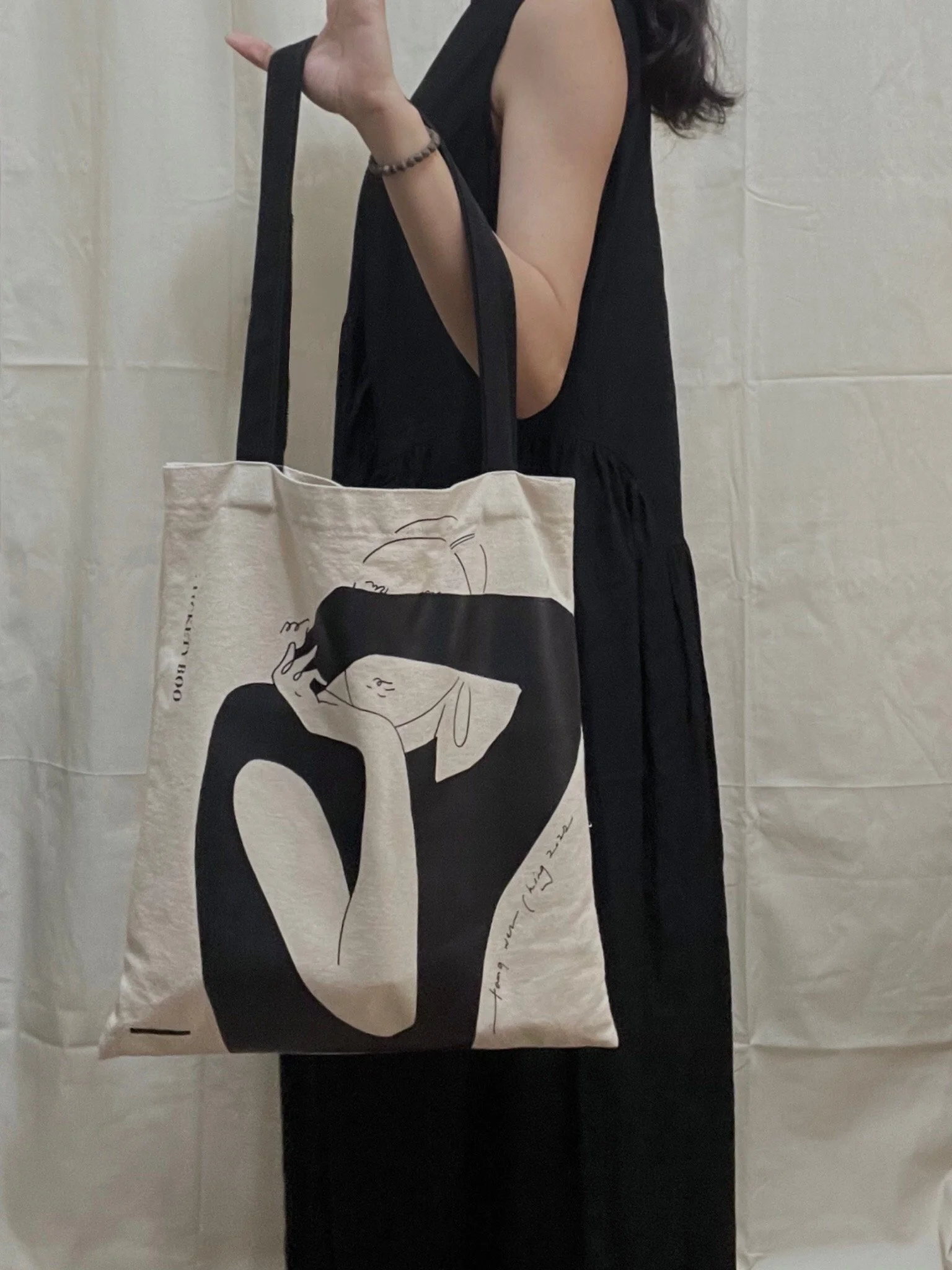 A beige tote bag with a black line drawing of a woman's face, held by a person wearing black dress and a black bracelet, standing in front of a plain white wall.