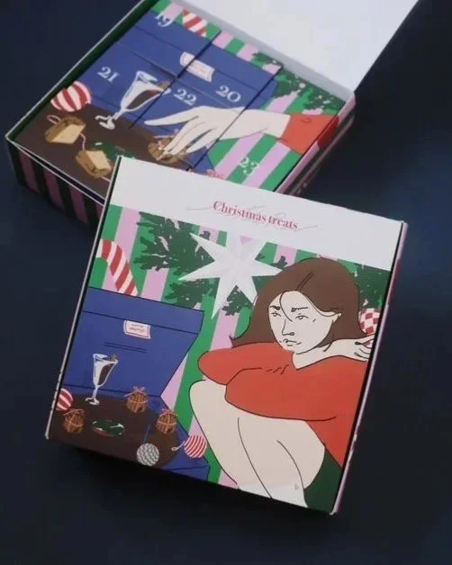 A decorated Christmas-themed advent calendar box with numbered doors and an illustrated woman resting her chin on her hand, surrounded by holiday items like gifts, ornaments, and a Christmas tree in the background.