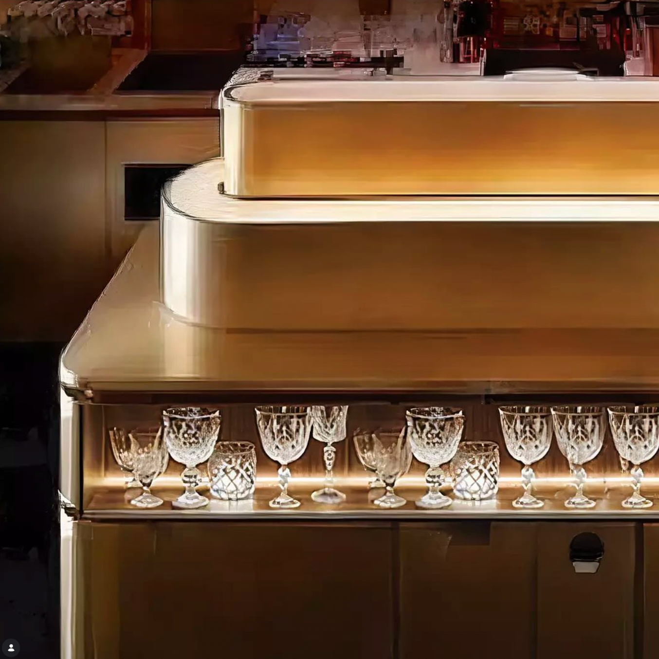 Golden island bar with a central bottle display and a collection of illuminated vintage cocktail glasses displayed on a shelf below.