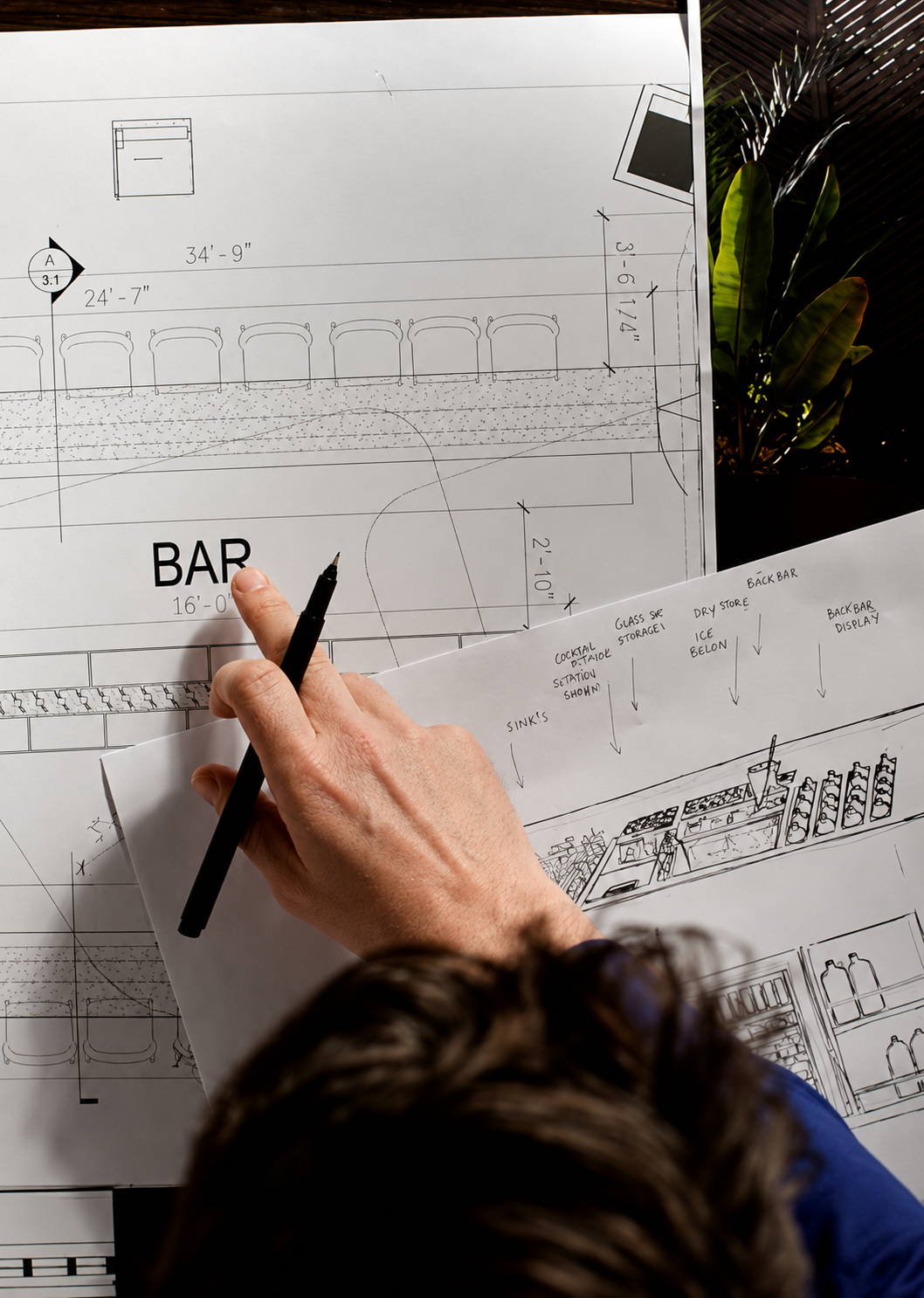 Bar designer drawing a measured bar layout on architectural plans for a new hospitality venue, with a hand holding a pen and a plant in the background.