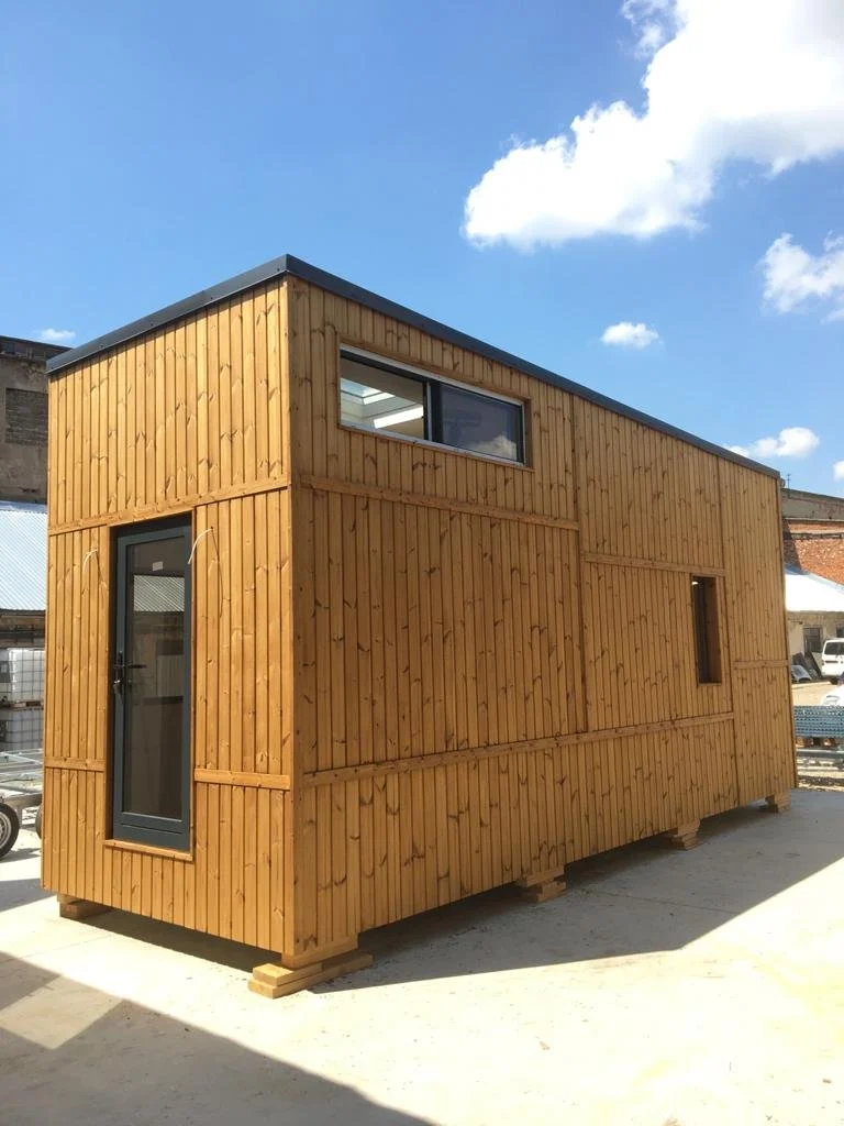A small, modern wooden cabin with a flat roof and large windows, raised on wooden blocks outdoors on a sunny day.