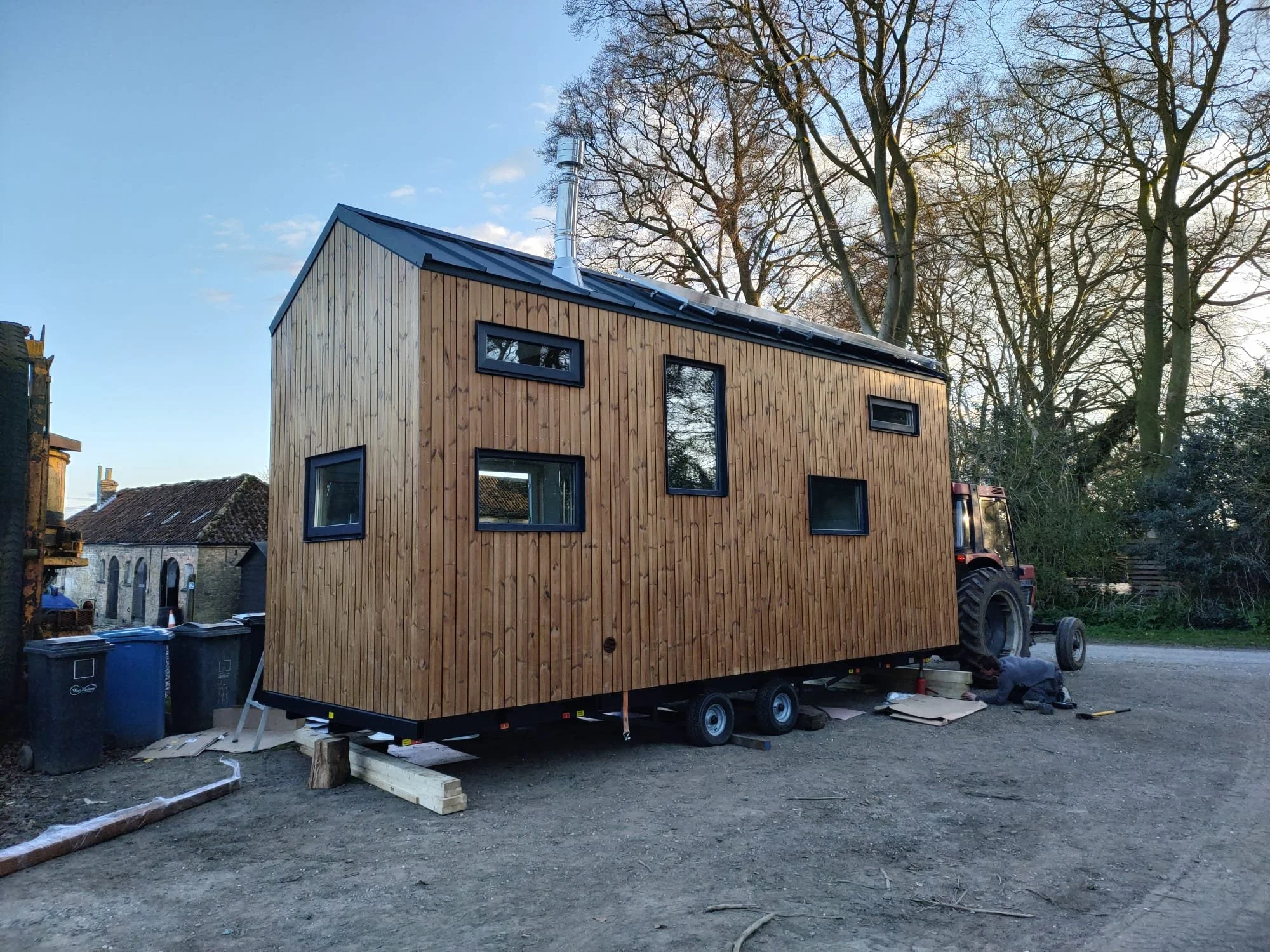 A tiny house or modular building with wooden siding, multiple irregularly placed windows, on a trailer attached to a tractor, outdoors with trees in the background.