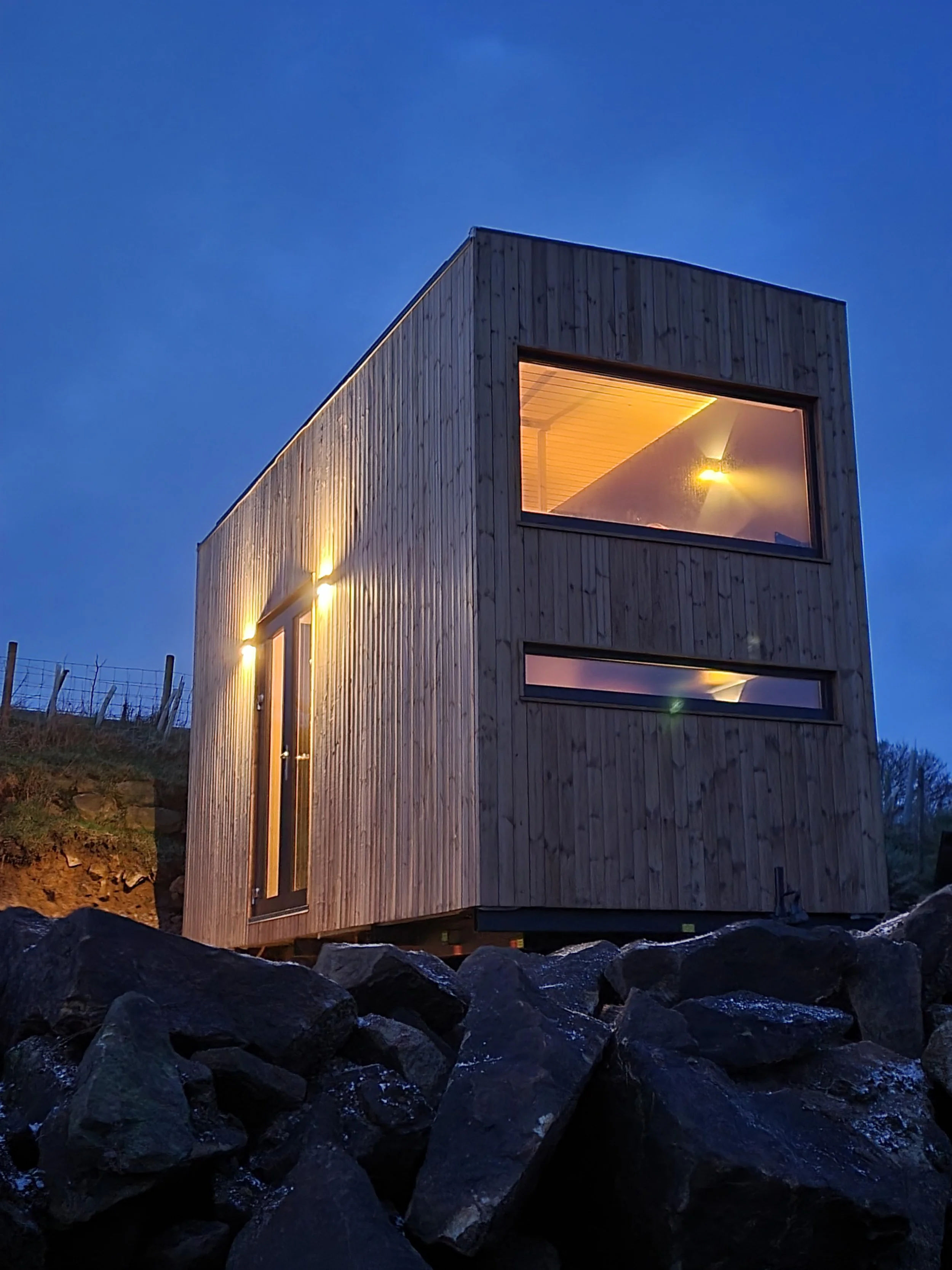 A modern two-story wooden house with large windows, illuminated from within, situated on a rocky landscape during dusk.