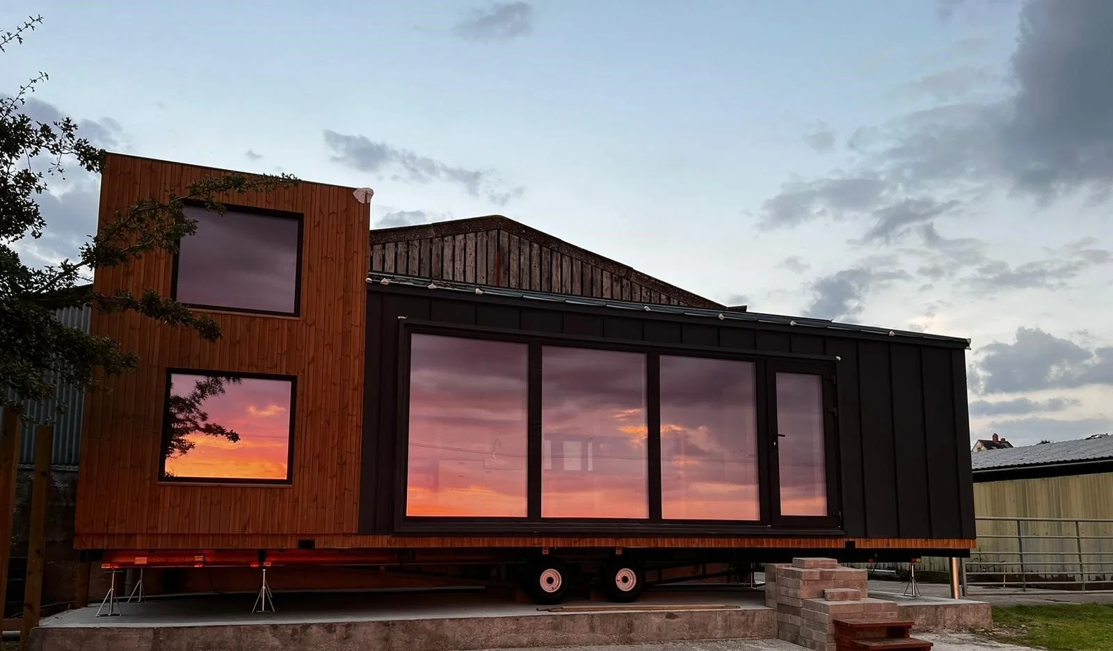 Modern tiny house with black and wood exterior, large windows reflecting a sunset, on a raised concrete foundation, with steps leading up to the entrance.