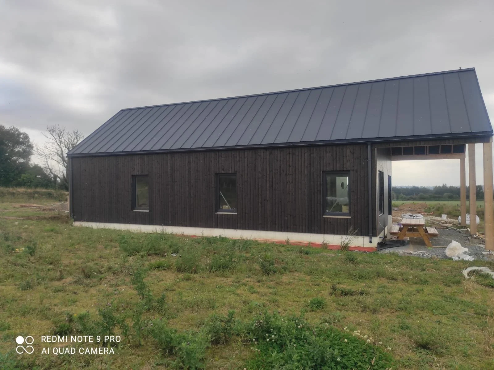 A black wooden house under construction on a grassy field with a gray overcast sky.