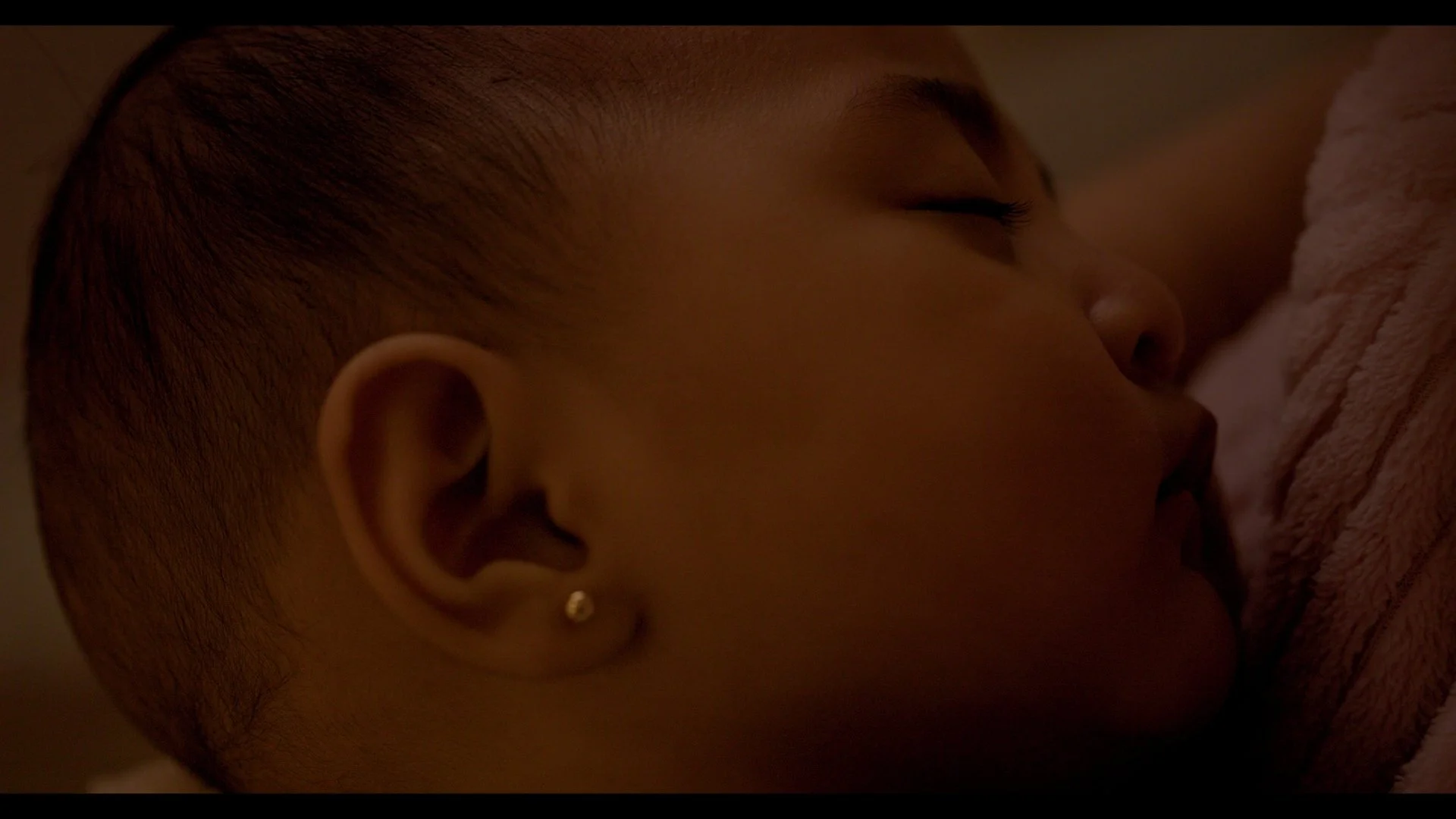 Close-up of a young person sleeping on a soft pink blanket, with short hair and a small earring.