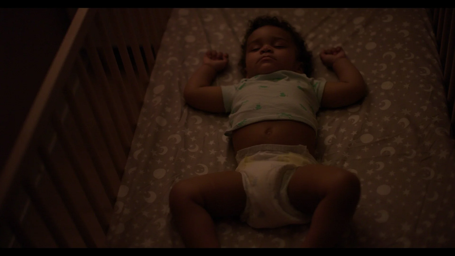 A young child lying on a bed in a crib with stars and moon pattern bedding, wearing a light-colored shirt and diaper, with arms raised.