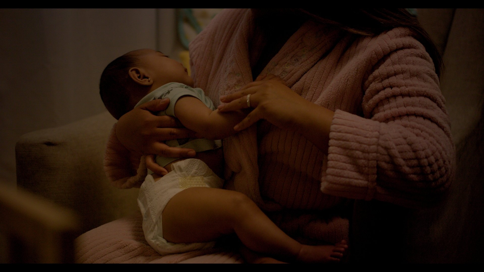 A woman wearing a pink sweater gently holding a sleeping baby on her chest in a dimly lit room.